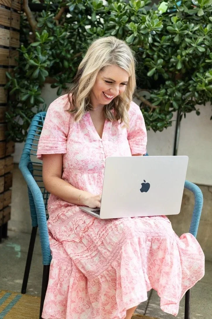 A woman with blonde hair sitting outside on a blue chair, smiling at a silver laptop with an Apple logo, wearing a pink dress with a floral pattern and a gold bracelet, surrounded by green foliage.