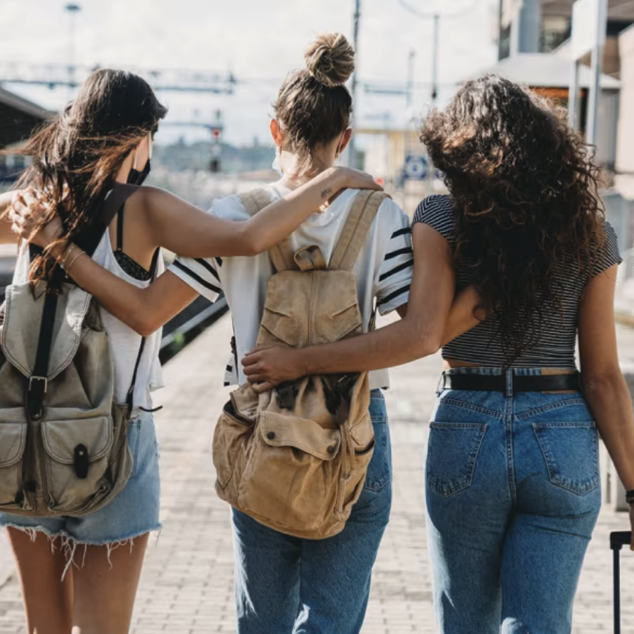 Three women walking together at a train station, with one woman resting her arm on the shoulders of the other two as they walk.