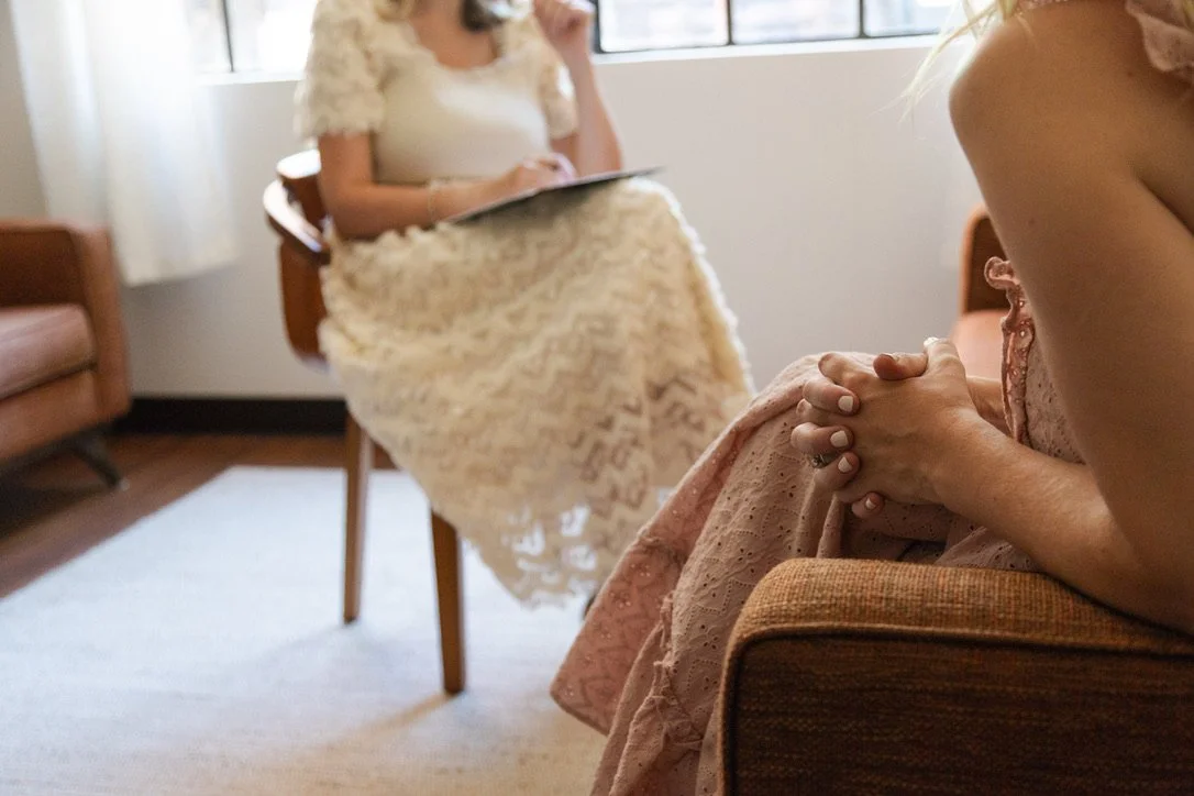 Two women seated indoors, one in the foreground with hands clasped, and one in the background wearing a cream lace dress, holding a tablet or notebook on her lap.