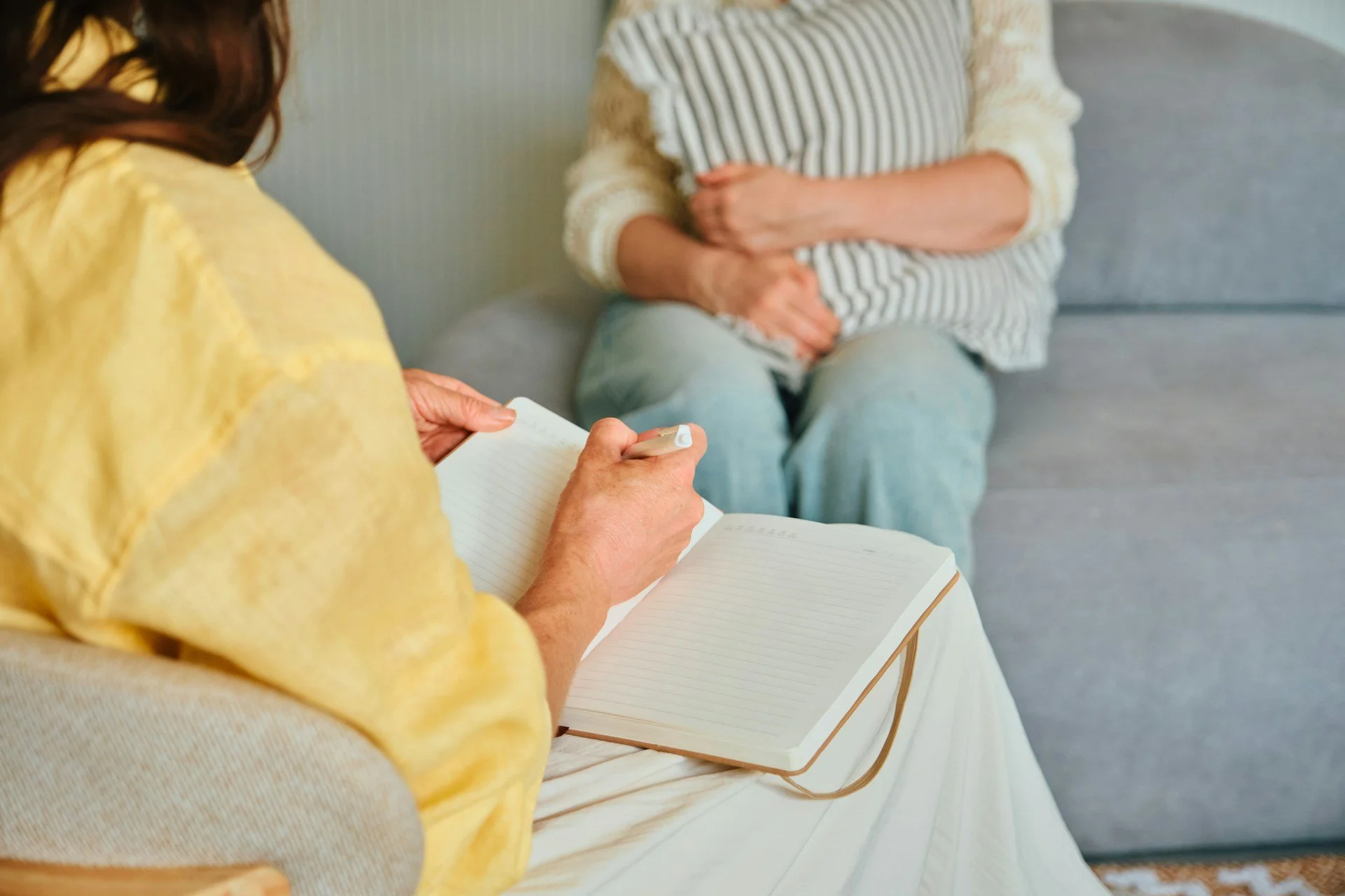 A woman in a yellow shirt writing in a notebook while sitting on a couch, with another person sitting nearby with their hands folded in their lap.