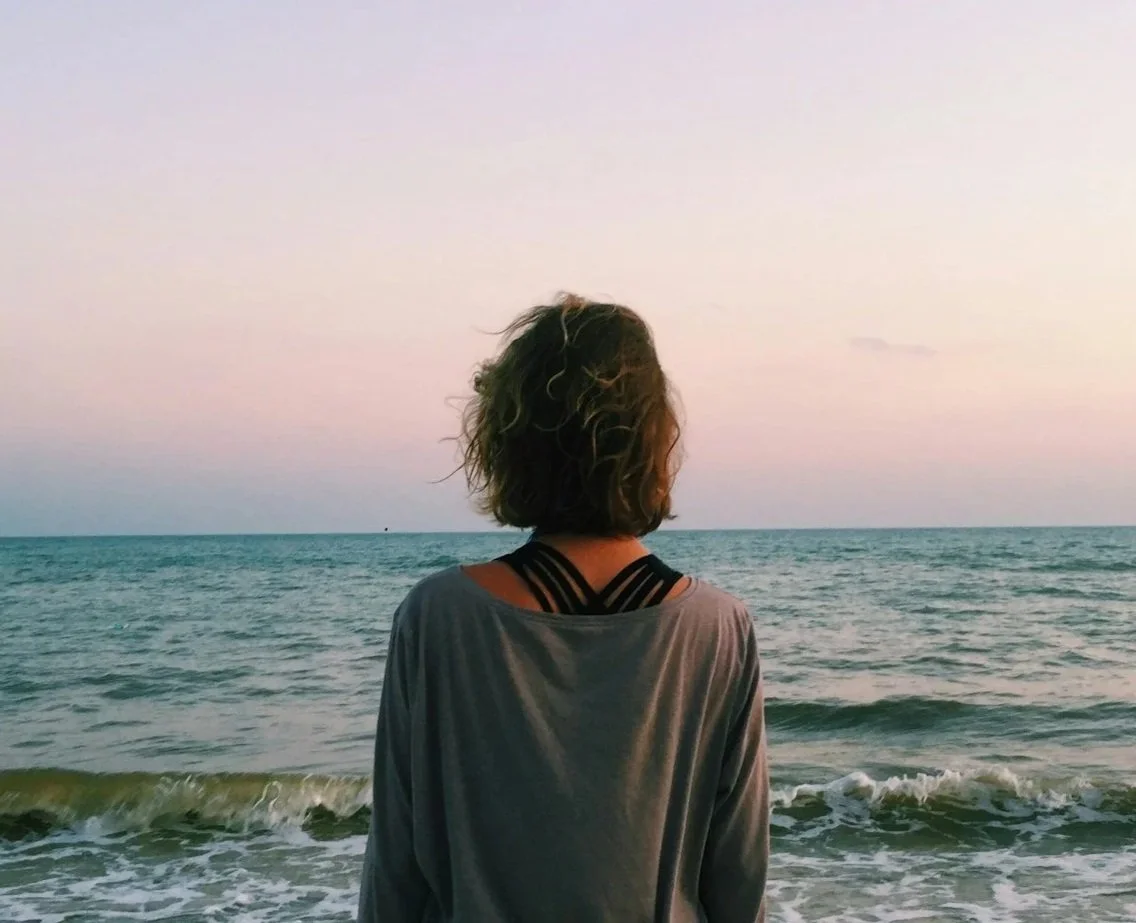 A person with curly hair facing the ocean at sunset, wearing a gray top and black strappy sports bra.