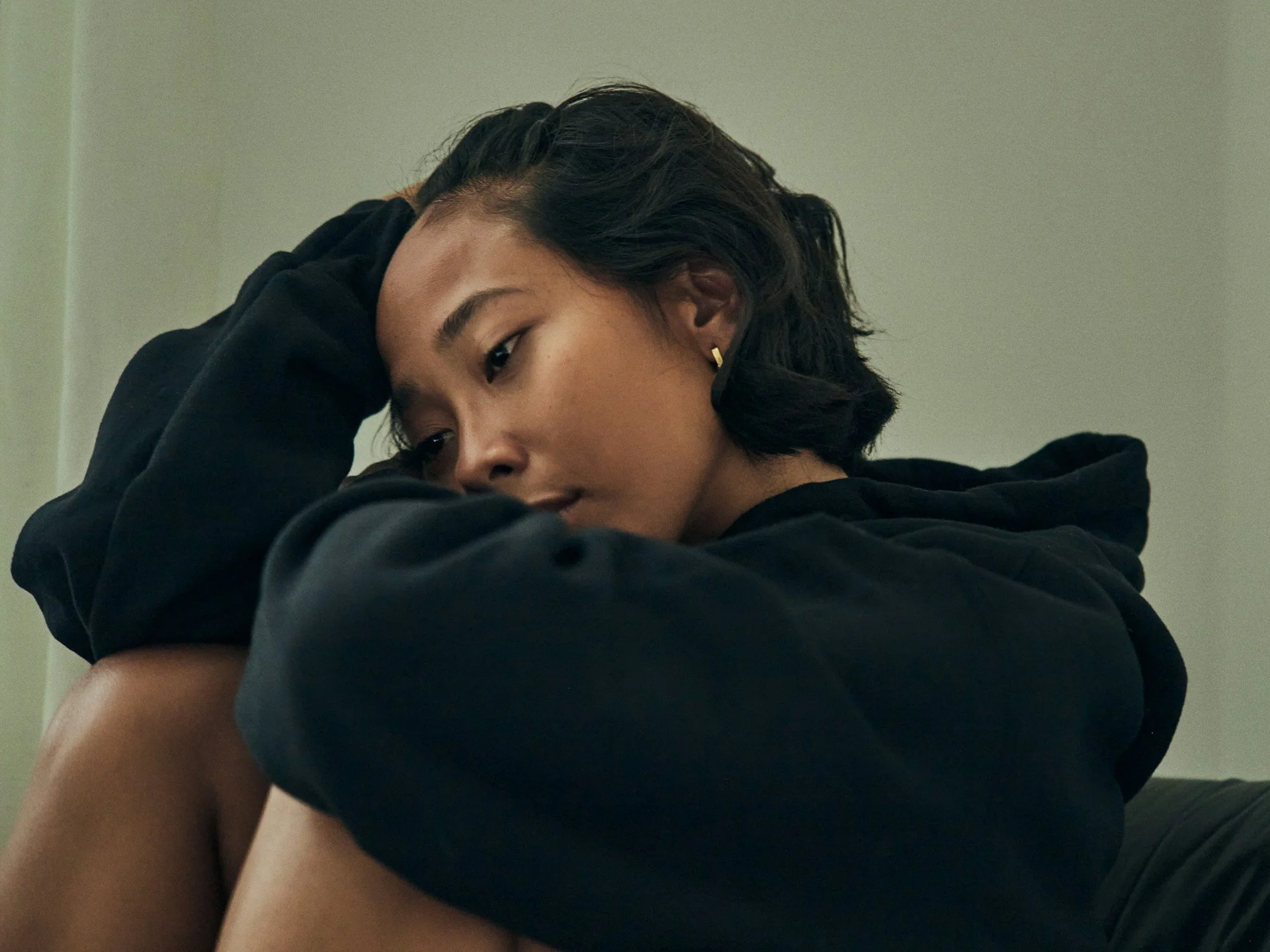 A woman with short dark hair wearing earrings, resting her head on her hand, dressed in a black hoodie, sitting with arms crossed on her knees against a plain wall background.