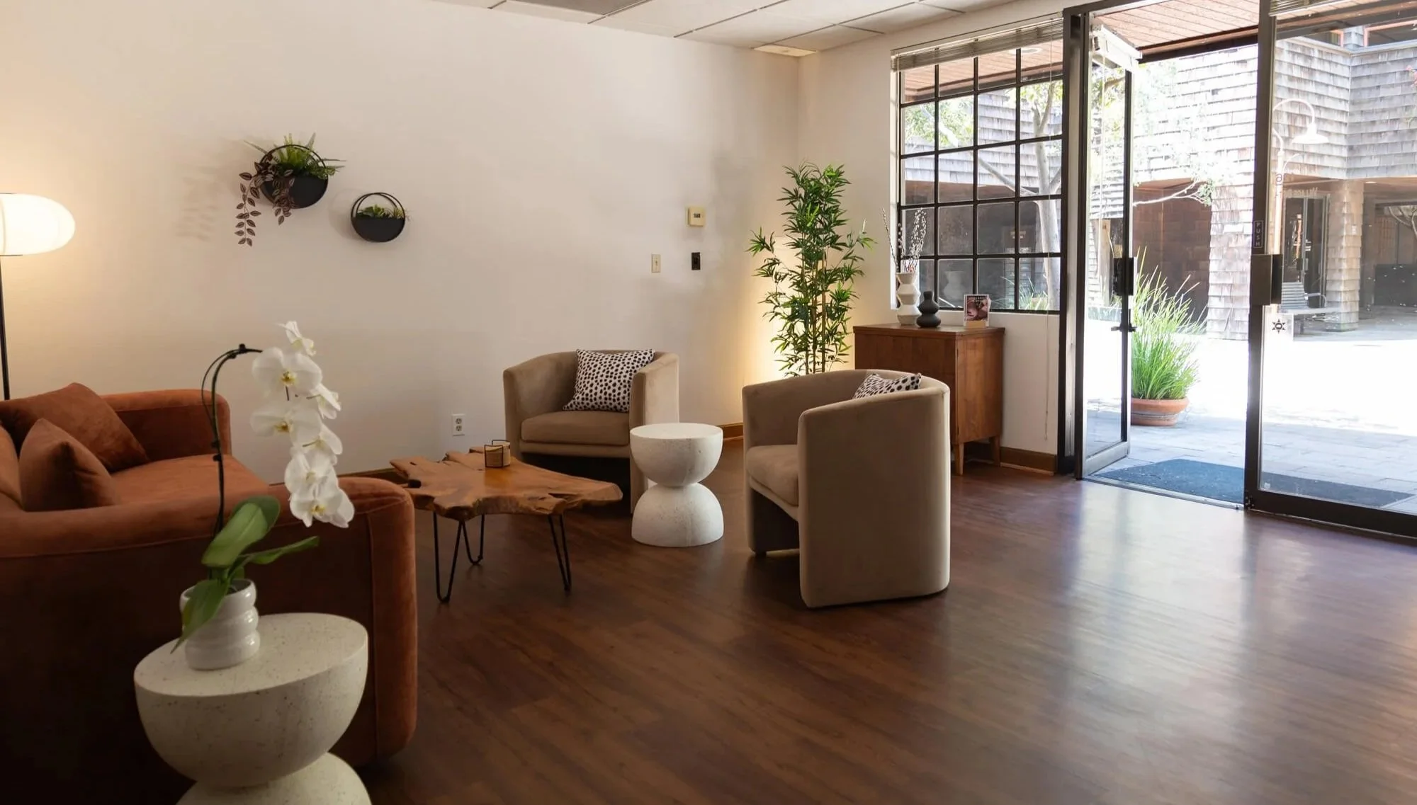 Interior of a modern living room with a brown sofa, beige armchairs, a wooden coffee table with a live edge, and various decorative items. There are plants on the walls and a large window and glass door letting in natural light.