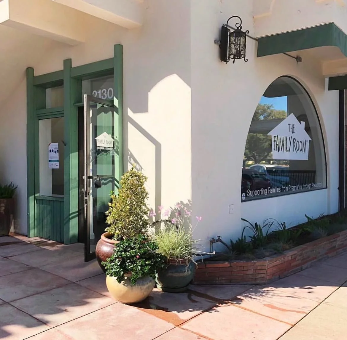 Exterior of The Family Room with a green door and large window, potted plants outside, and a sign supporting families from pregnancy through parenthood.