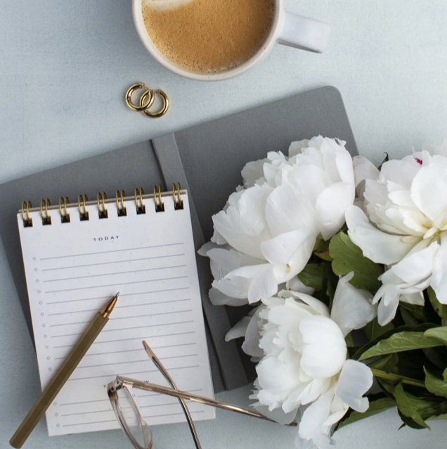 A white notebook with a gold pen, a pair of reading glasses, a cup of coffee, a bouquet of white flowers, and two gold rings on a light blue surface.