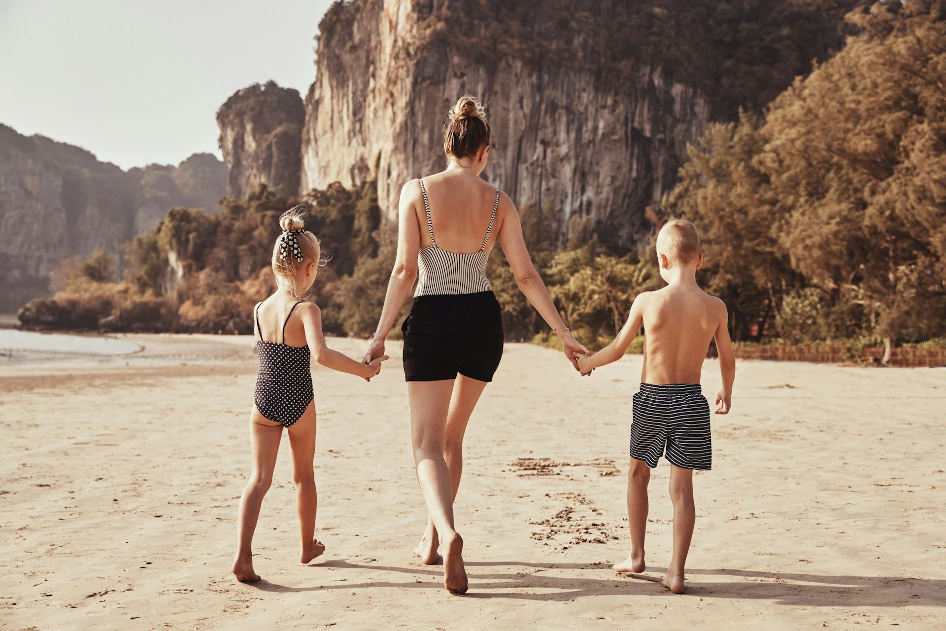 Woman walking on beach with two young children, holding hands, with rocky cliffs and trees in the background.