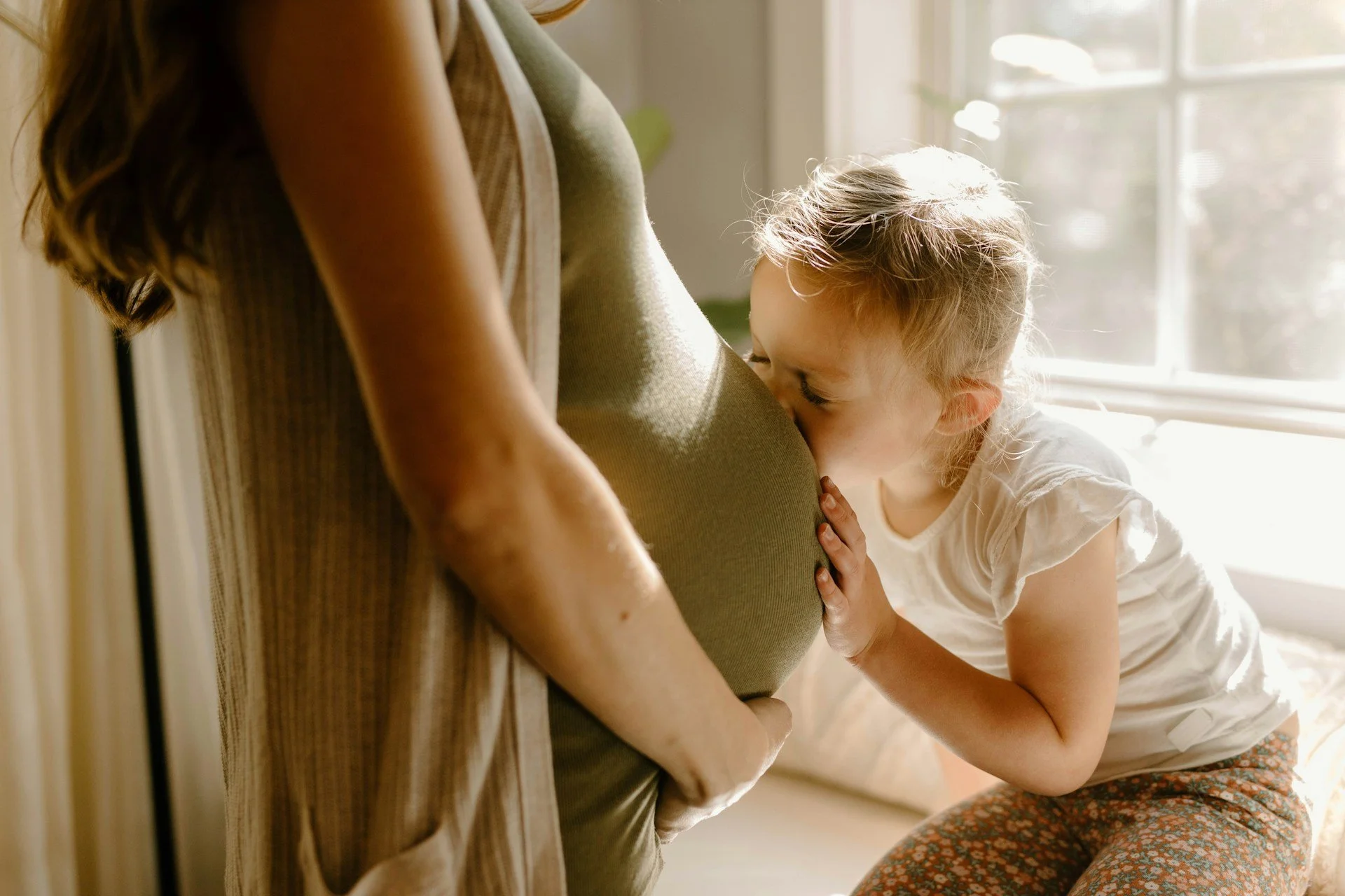 A young girl hugging a pregnant woman, resting her head on the woman's belly as the woman stands by a sunlit window.