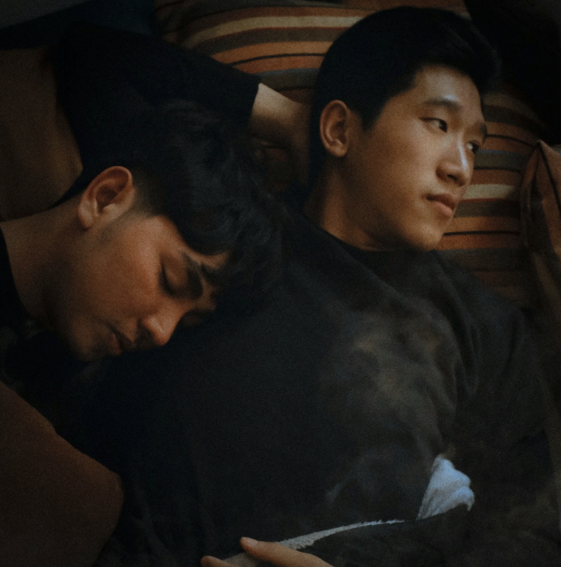 Two young men lying on a striped couch, one resting and the other looking away.