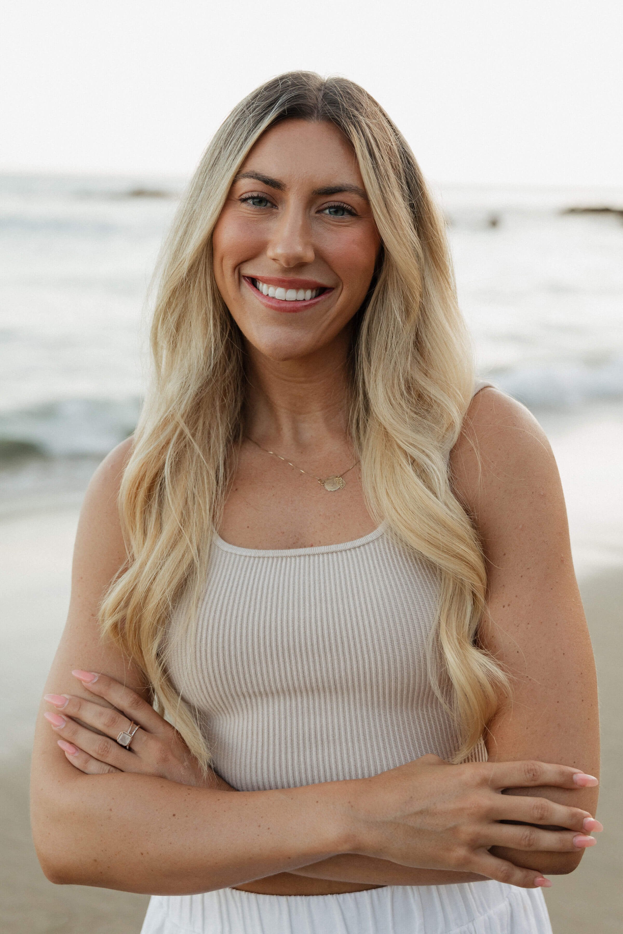 A smiling woman with long blond hair standing on the beach, wearing a white tank top and crossing her arms.