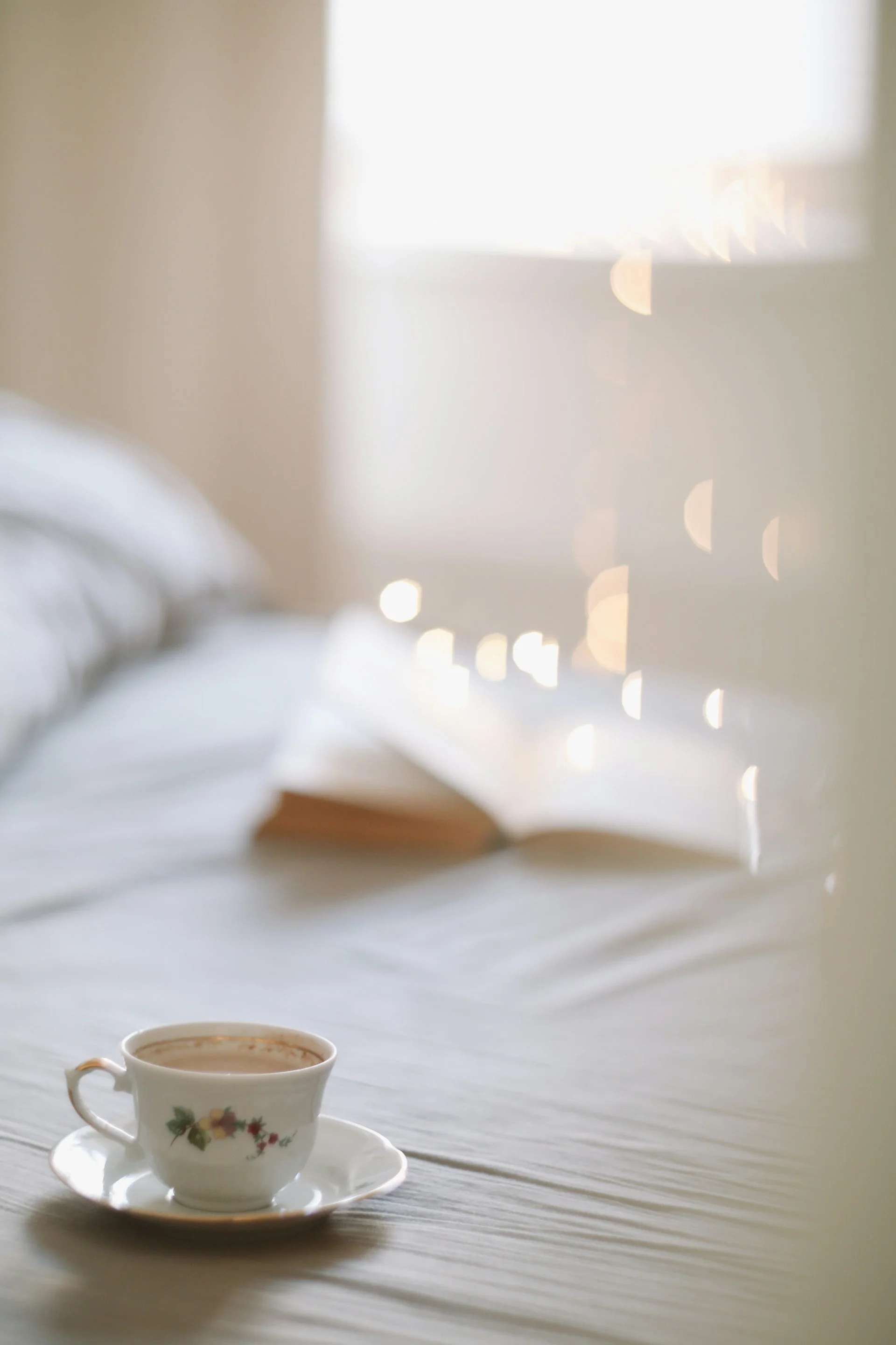 A white teacup with floral design on a matching saucer on a light-colored table. The background shows a bright window with sunlight and small bokeh light spots.