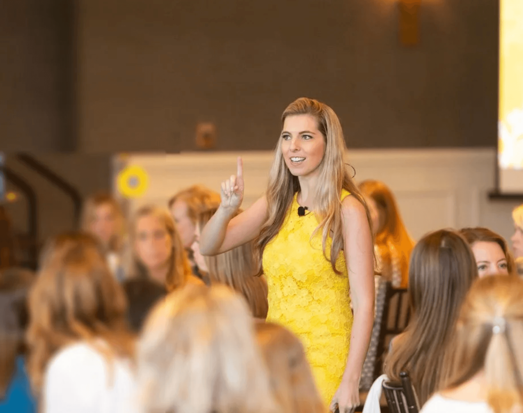 A woman in a yellow dress stands with her mouth open, raising her right hand with her index finger up, addressing a group of young women seated in a room with brown walls and chairs.