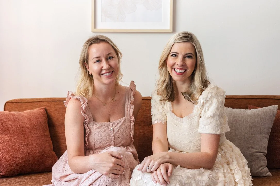 Two women sitting on a brown couch smiling at the camera, with a framed picture on the wall behind them.