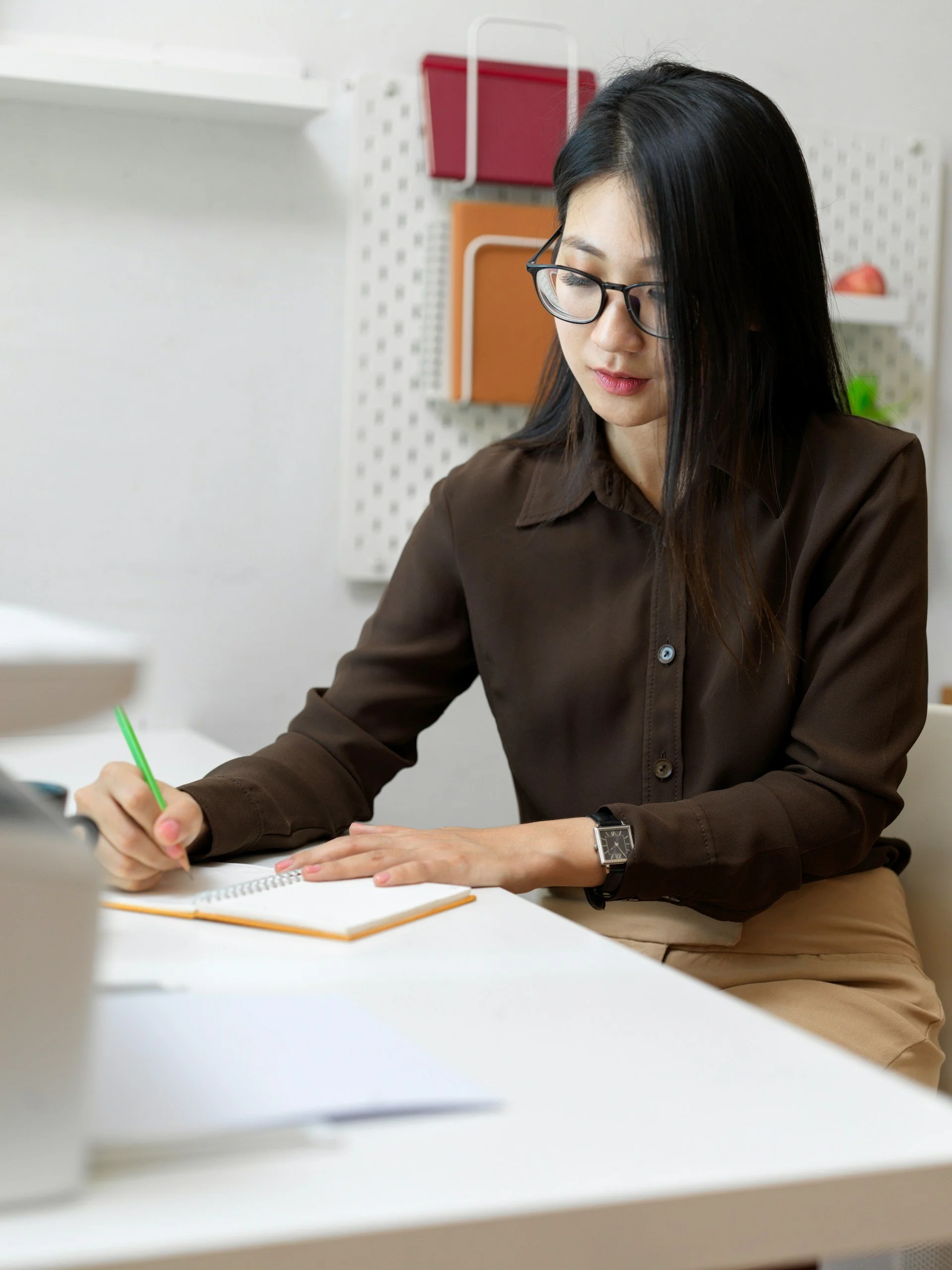 A woman with long black hair and glasses, wearing a brown shirt, is sitting at a white desk writing in a notebook with a green pen. There is a smart watch on her left wrist and a laptop partially visible on the left side of the foreground. In the background, there is a white wall with a pegboard, colorful folders, and a small red object.