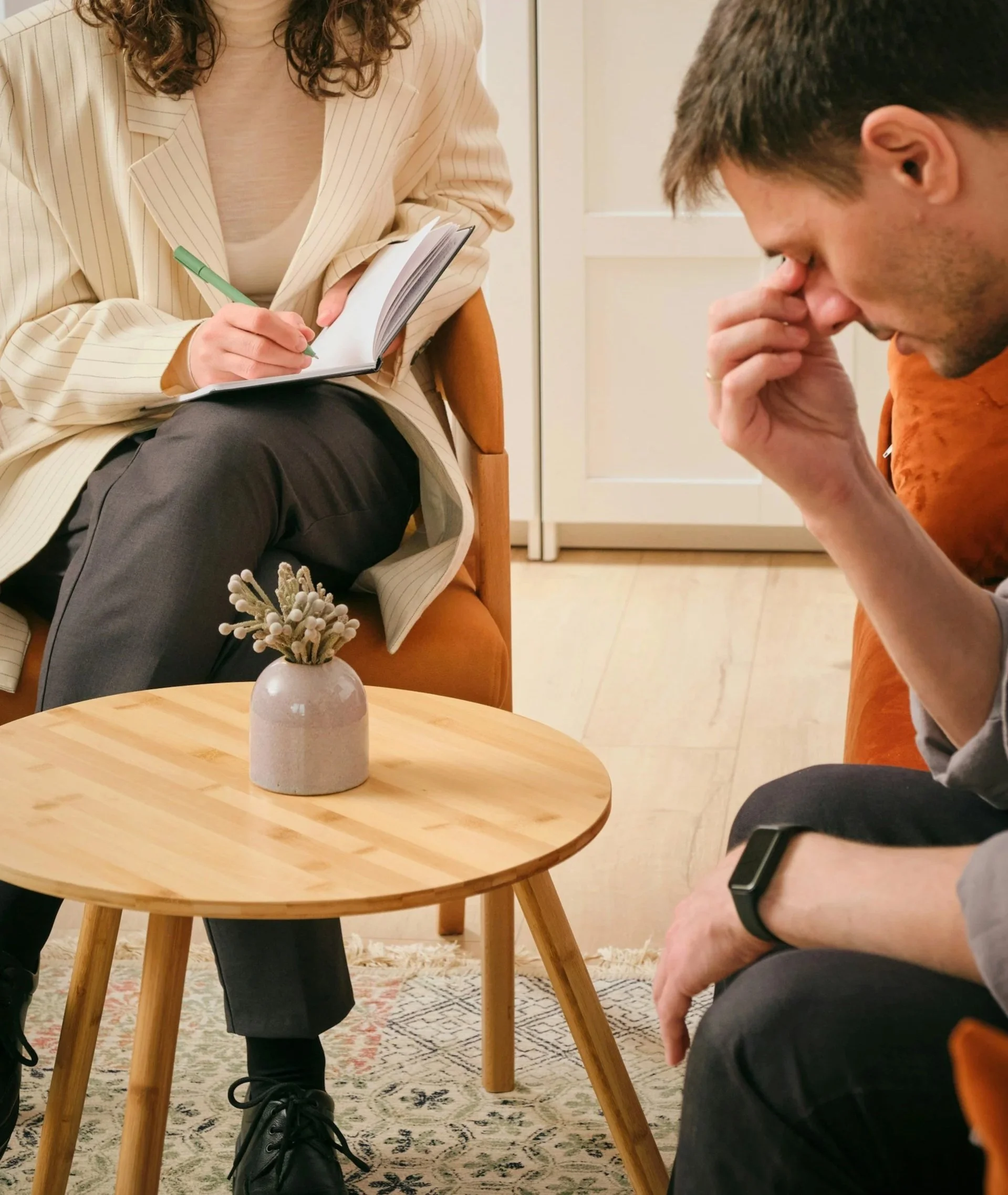 A woman seated at a wooden table taking notes with a green pen, and a man sitting nearby with his head bowed and hand on his face, in an indoor setting with a beige door in the background.