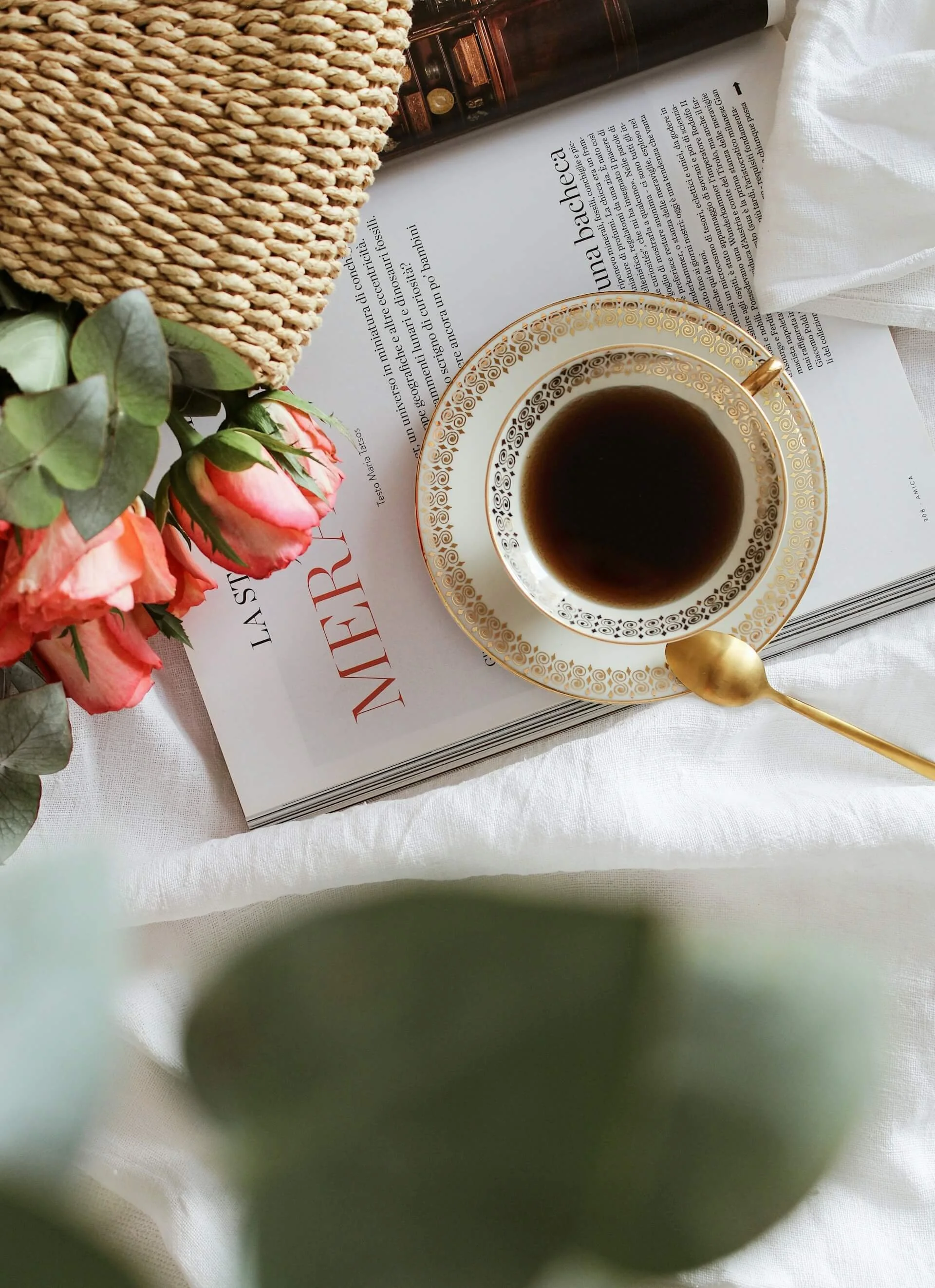 A cup of black coffee in a white and gold ornate cup and saucer, placed on top of a magazine or book on a white cloth surface, with pink roses and green leaves, and a woven basket and a brown bottle in the background.