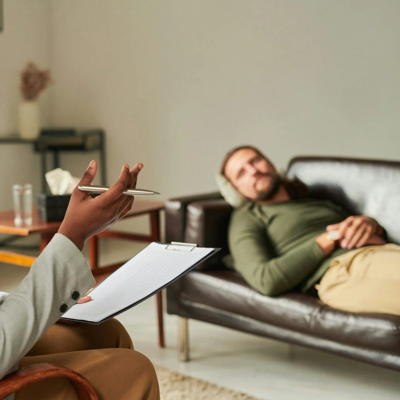A man lying on a sofa with a green shirt and beige pants, wearing headphones, while a person in a beige blazer takes notes on a clipboard and holds a pen.