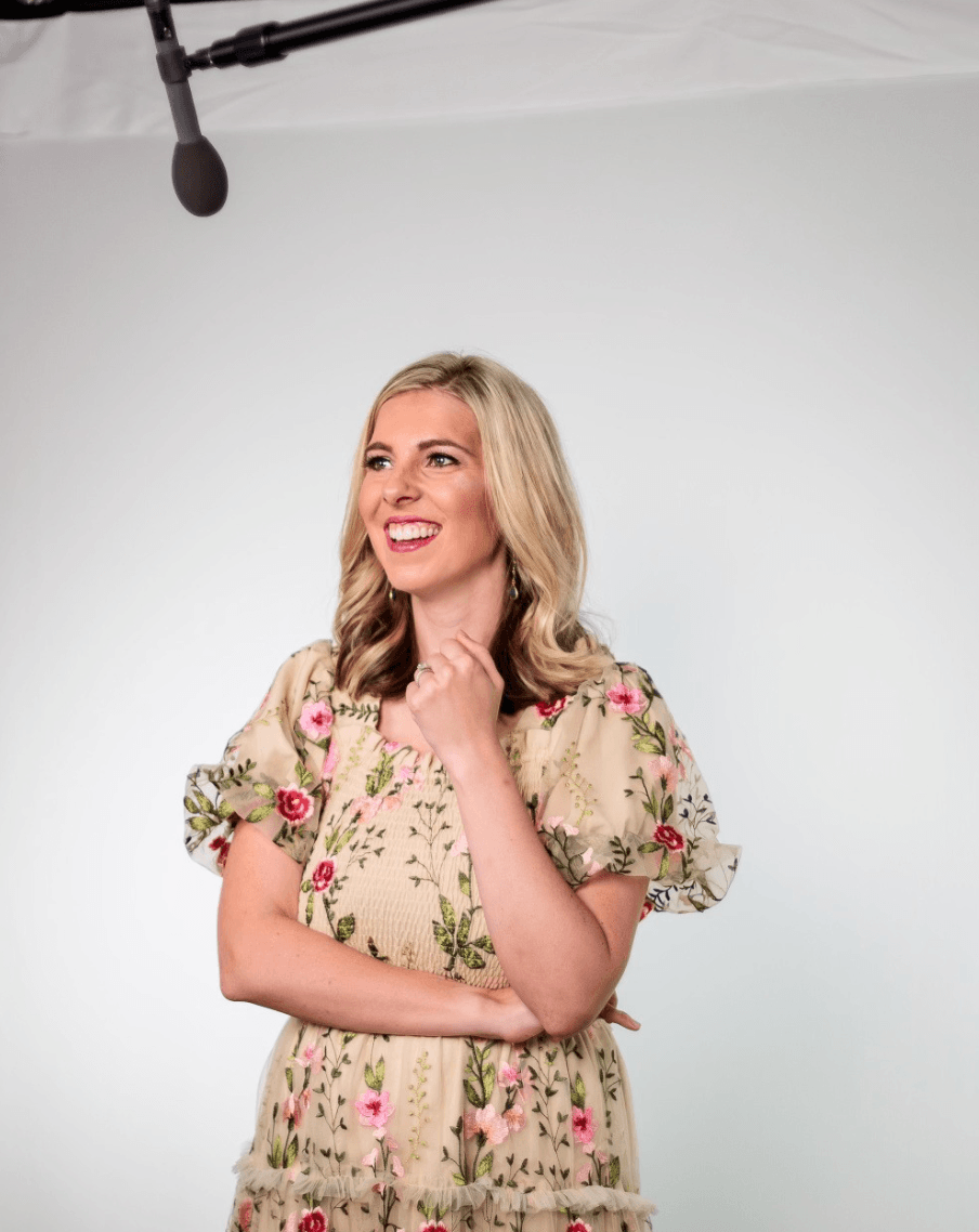 A woman with blonde hair smiling in a floral dress stands in front of a white background, with a microphone hanging from the ceiling above her.