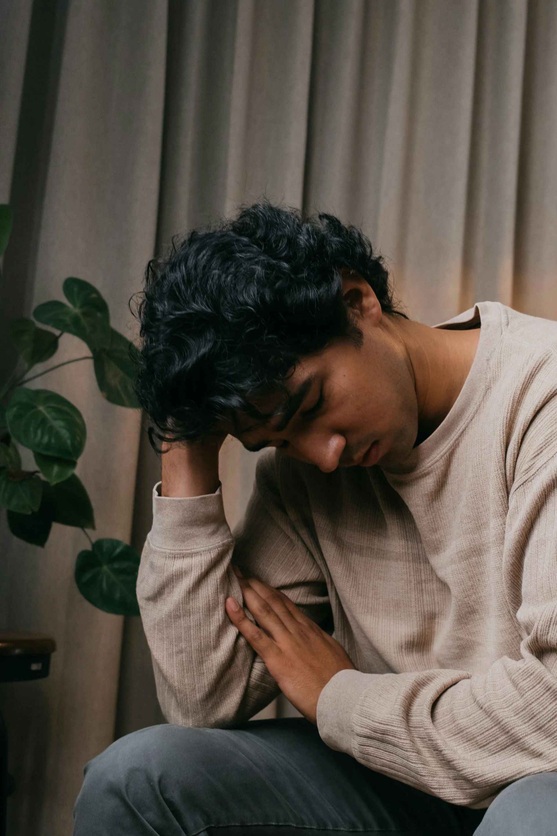 A person with dark curly hair sitting with their head resting on their hand, appearing to be upset or distressed, with a plant and beige curtains in the background.