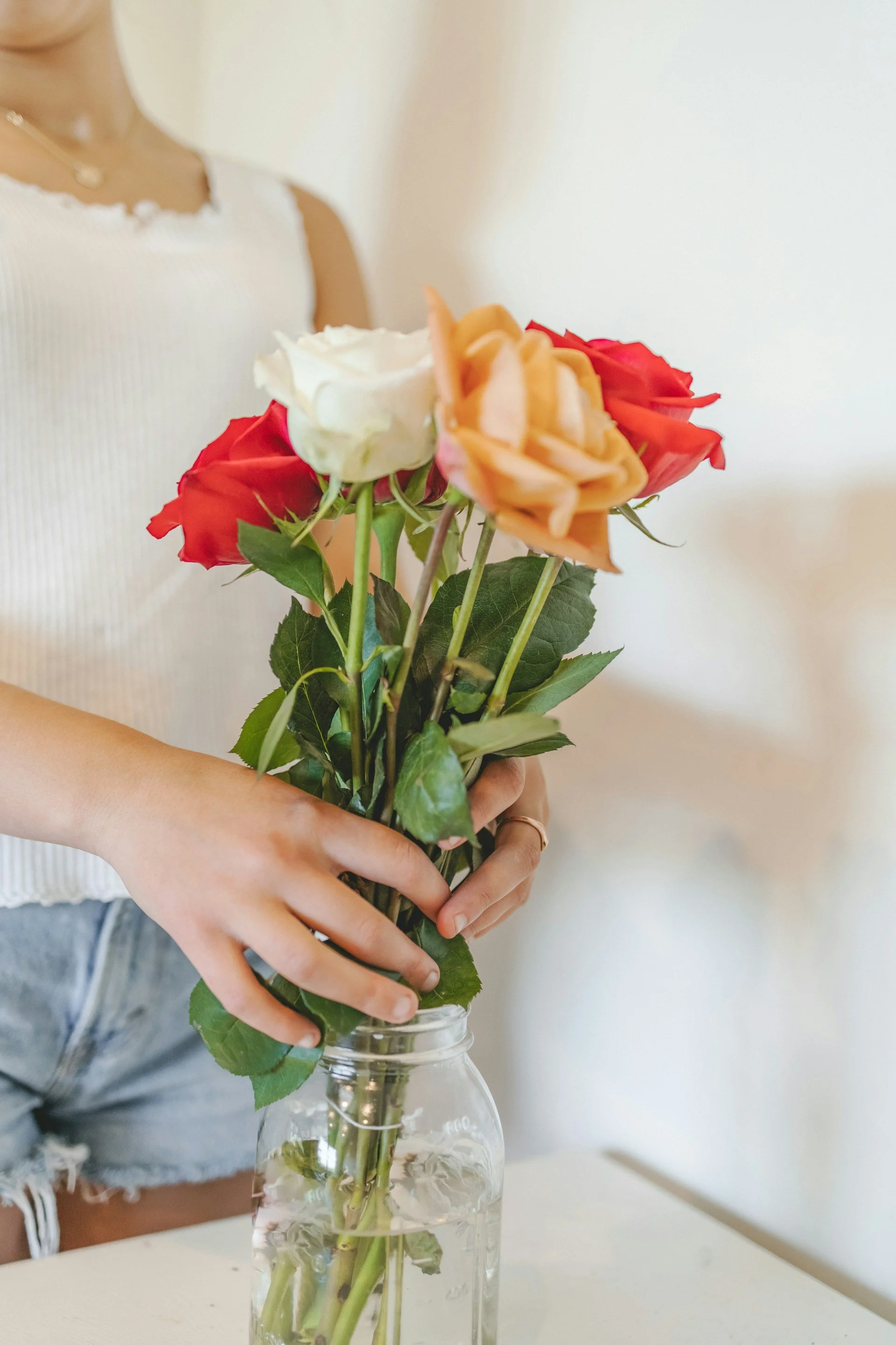 A person holding a bouquet of mixed color roses in a glass jar on a white table against a plain white wall.