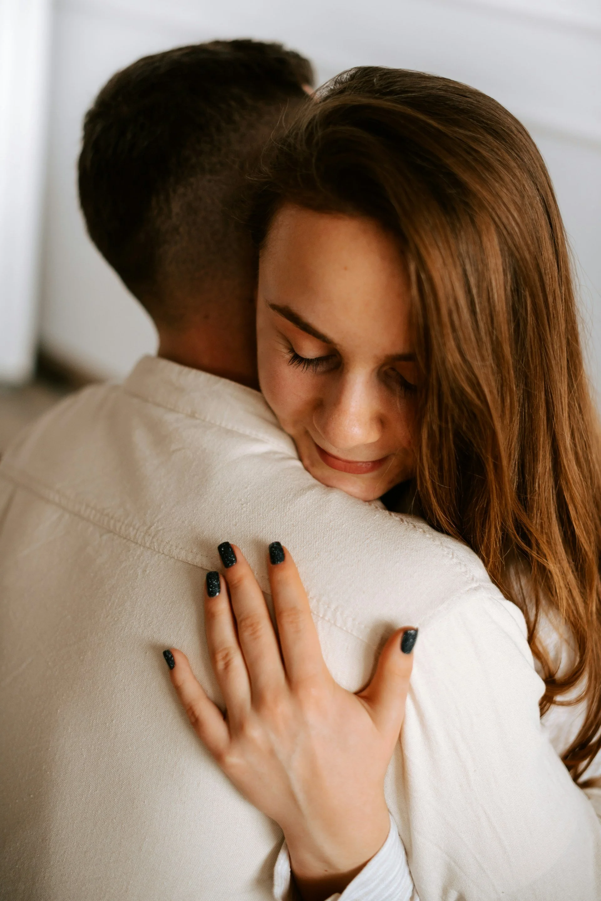 A woman with long red hair hugging a man with short dark hair, who is facing away. She has her eyes closed and is smiling softly, with her hand on his back.