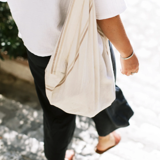 Person walking barefoot on a beach with a white tote bag over their shoulder, wearing a white top and black pants.
