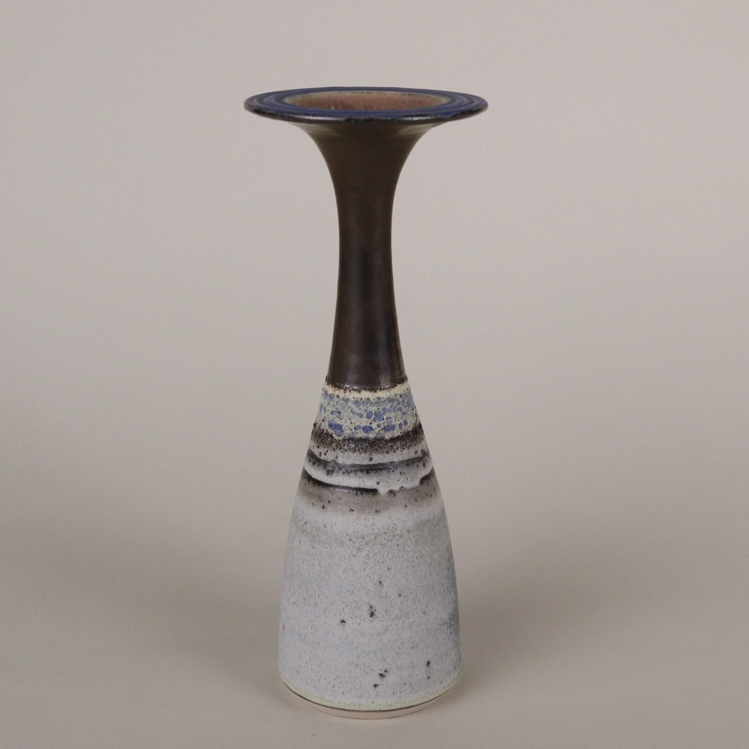 Ceramic vase handmade by Emma Baldwin with a slim, tapering form, featuring a bronze upper section, a transition with blue and white speckled glaze, and a white mottled lower section with black speckles, set against a plain background.