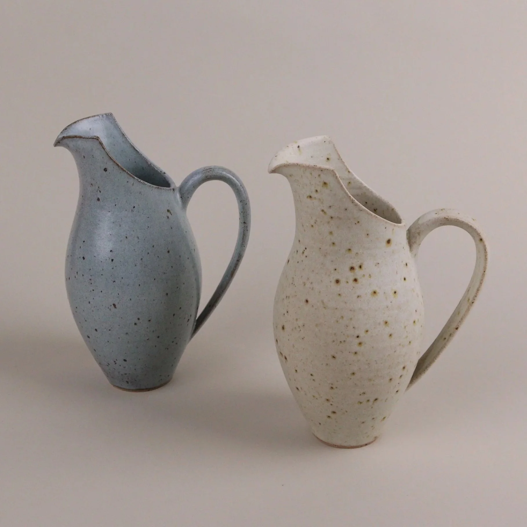 Two ceramic pitchers, one blue with speckles and one off-white with brown spots, on a plain background.
