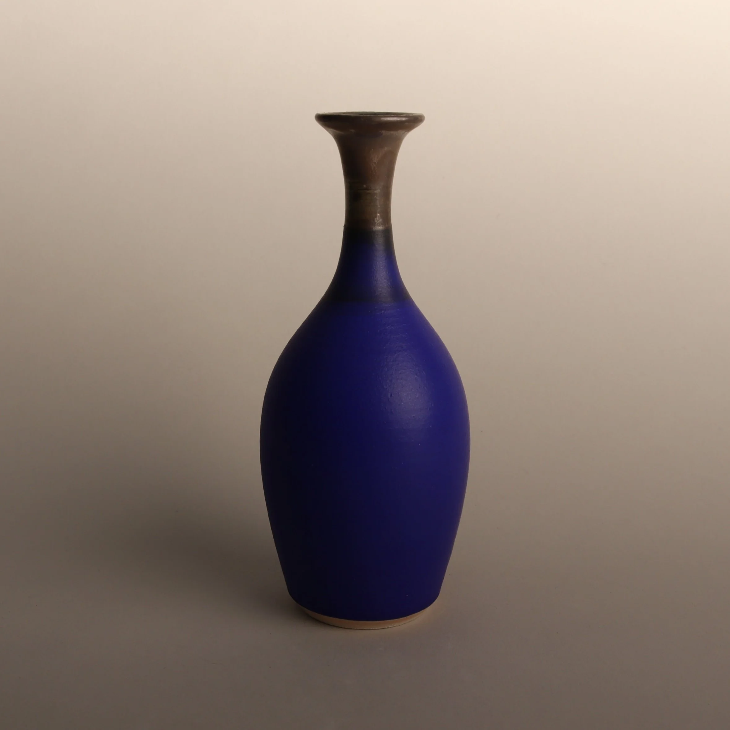 One of Emma Baldwin's ceramic bud vases with an intense matt blue body and a bronze neck, with a narrow neck and a flared opening set against a neutral background.