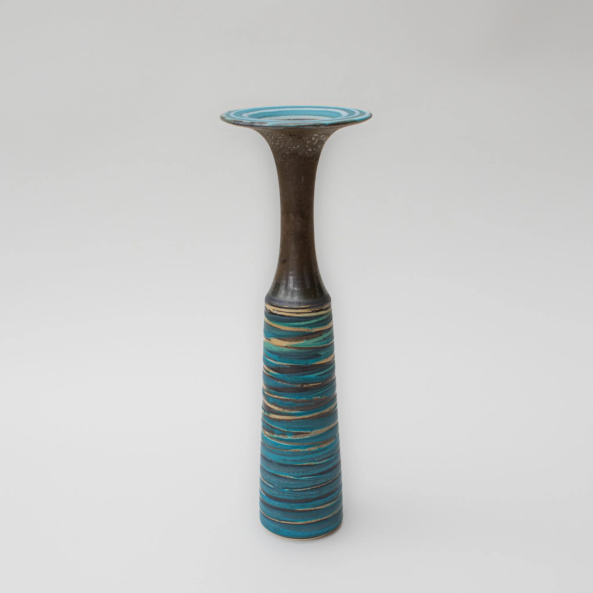 A tall, ceramic vase handmade by Emma Baldwin with a bronze glaze upper section and a turquoise, striped lower section, set against a plain white background.