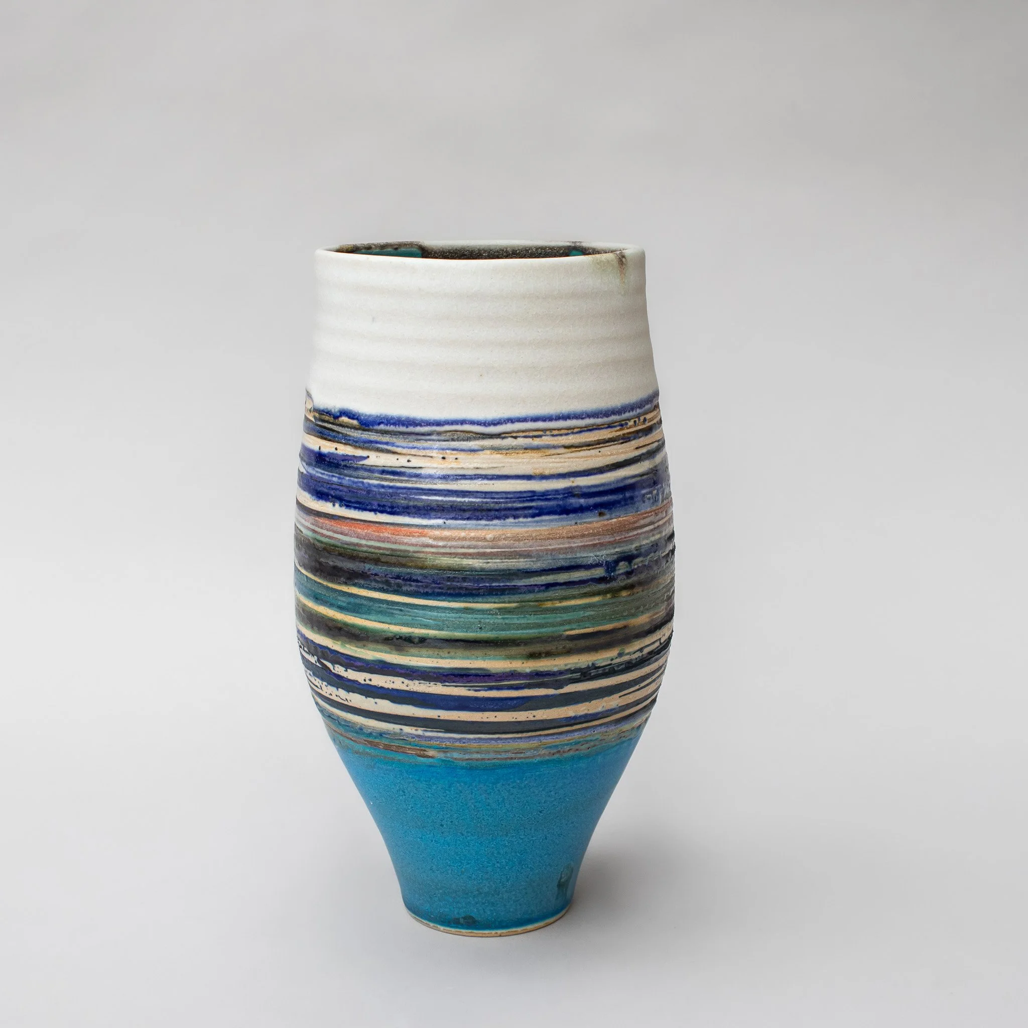 Ceramic vase handmade by Emma Baldwin with a white top and colorful horizontal stripes, transitioning from white to blue to turquoise, against a plain gray background.
