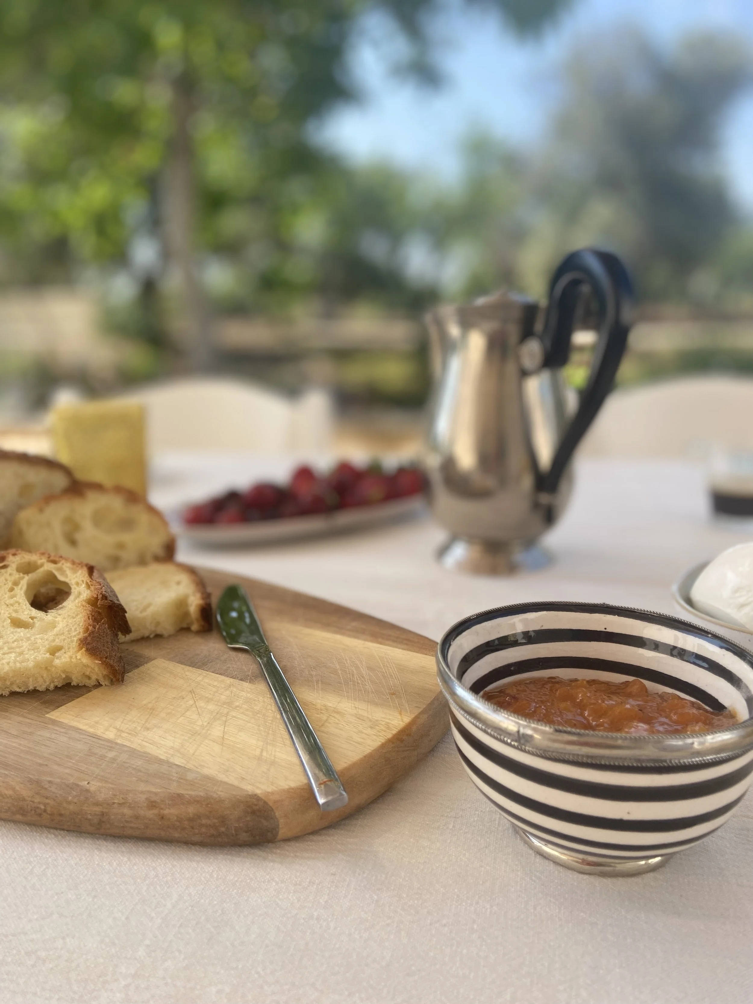 A breakfast table with sliced bread on a wooden cutting board, a small spoon, a bowl of jam, a pitcher, and a bowl of ice cream or yogurt, outdoors with trees and sky in the background.
