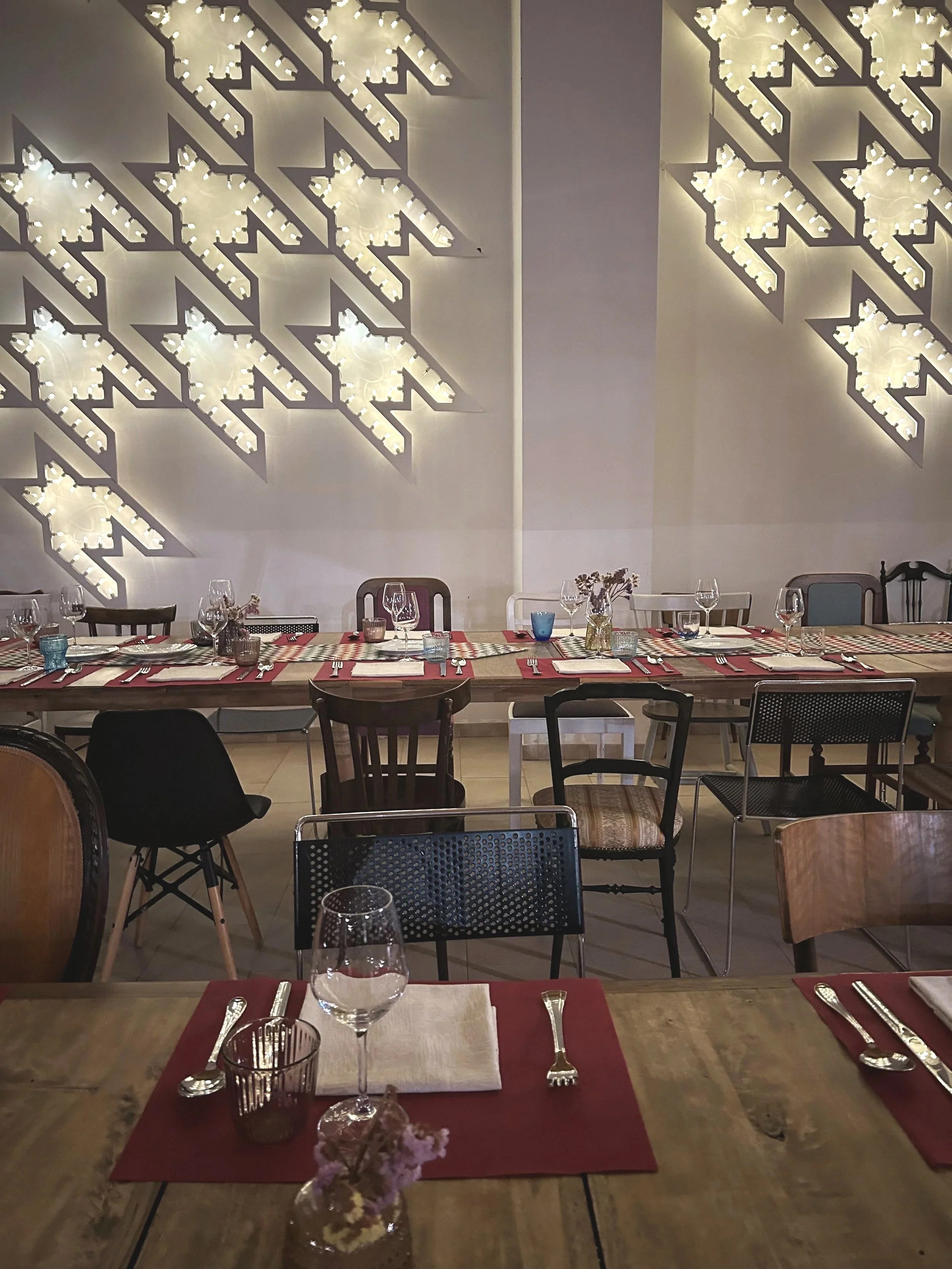 A dining room with a long wooden table set with wine glasses, utensils, and napkins, and a wall decorated with illuminated geometric light fixtures.