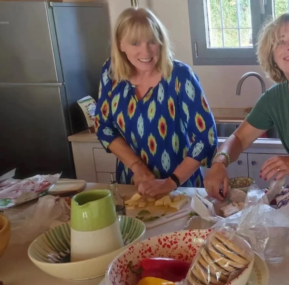A woman with blonde hair smiling in a kitchen, wearing a blue dress with a colorful pattern, standing at a countertop with food items and a woman in a green shirt next to her.