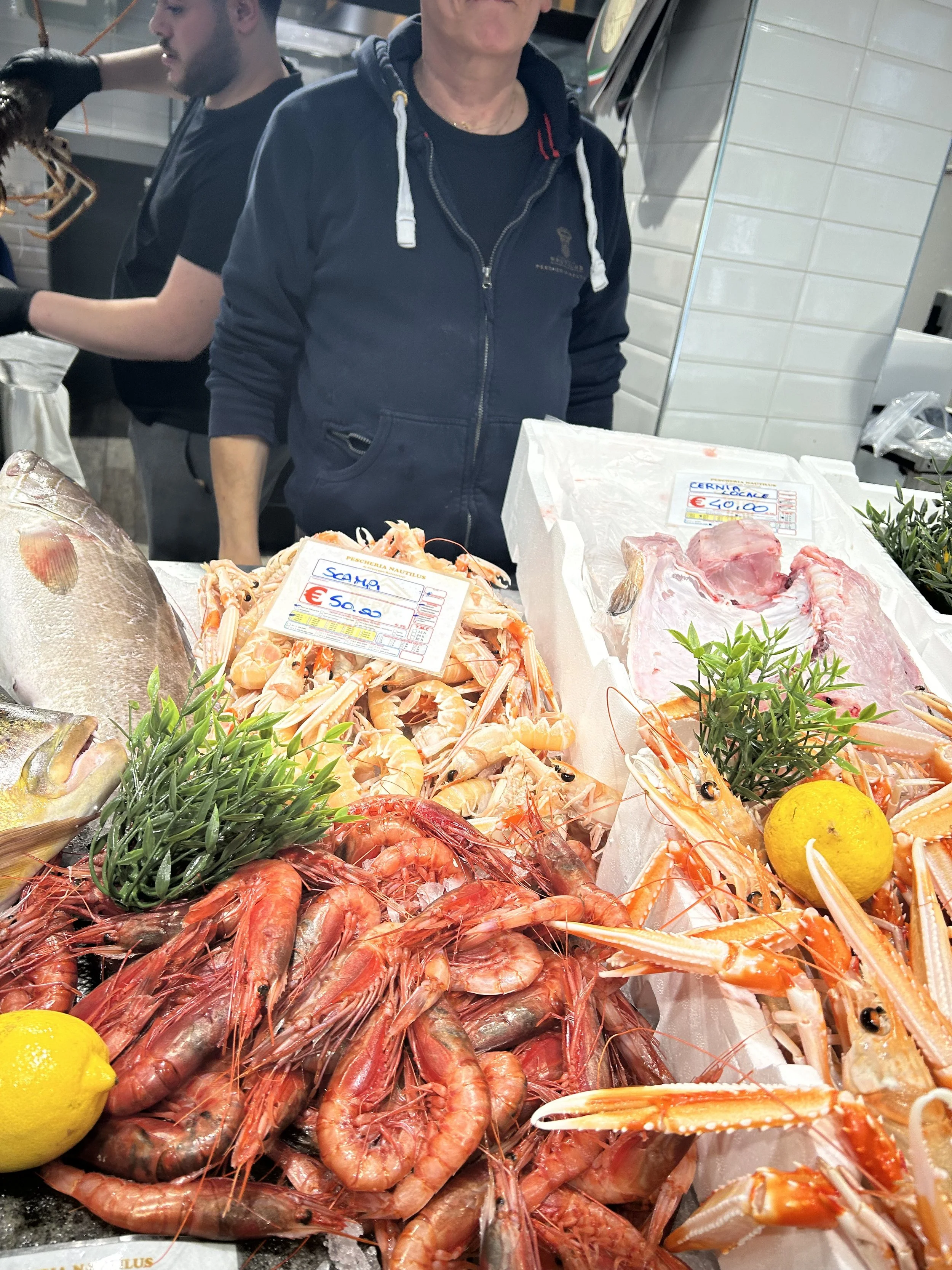 Fresh seafood display featuring shrimp, lobster, and fish at a market stall with two people in the background.