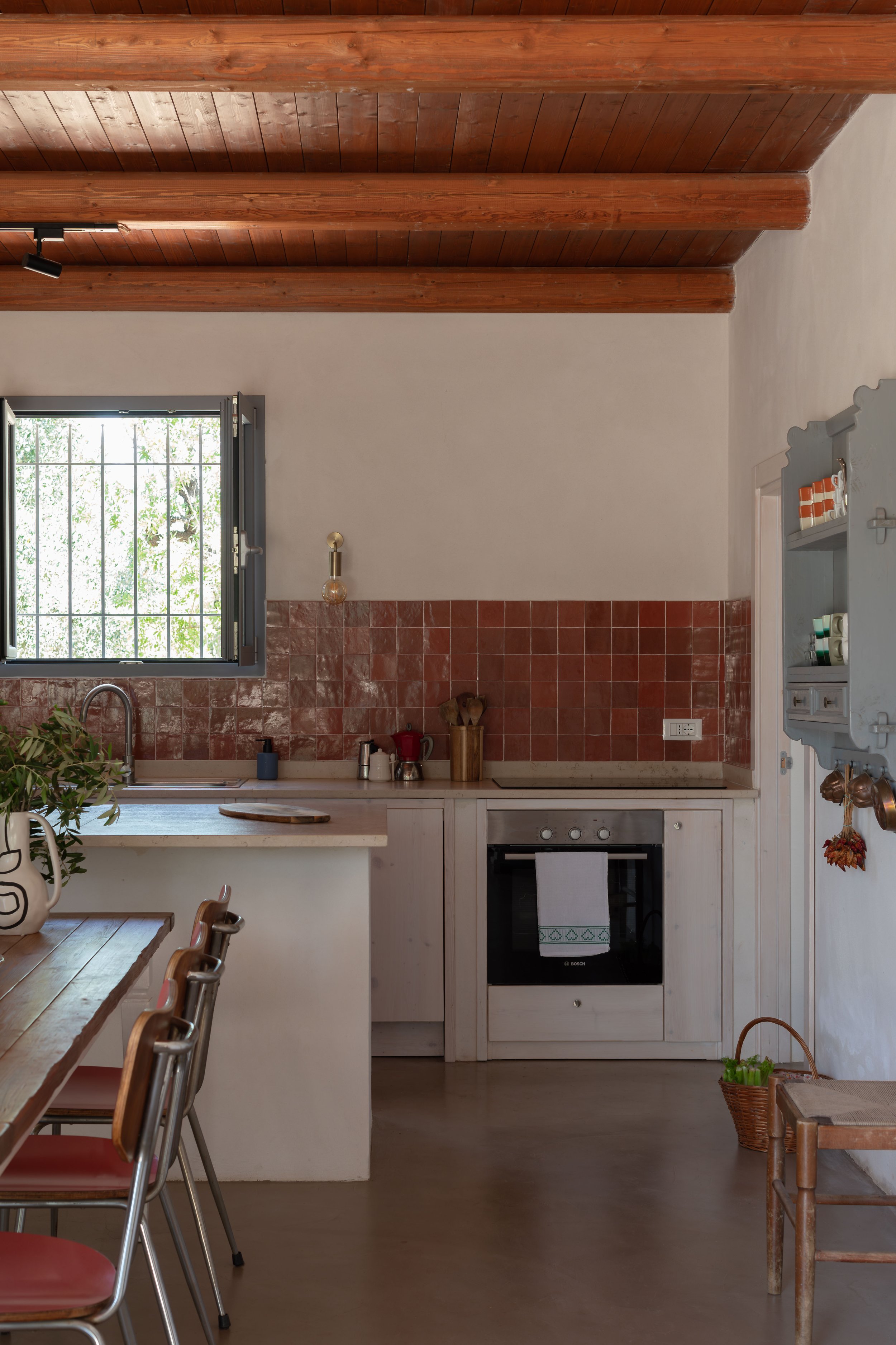 Kitchen with wooden ceiling beams, red tiled backsplash, window with bars, and white cabinets. There are various utensils and appliances, including an oven, sink, and a wooden table with chairs.