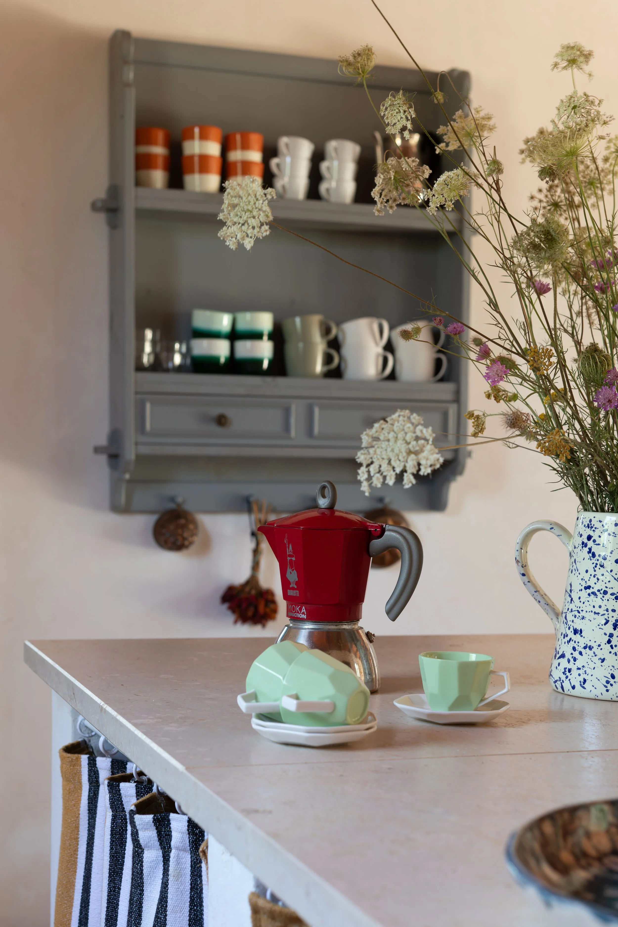 Kitchen with a grey wall cabinet, a red moka pot on the counter, with green and white cups on small plates, a large blue and white speckled pitcher, and a vase with assorted wildflowers.