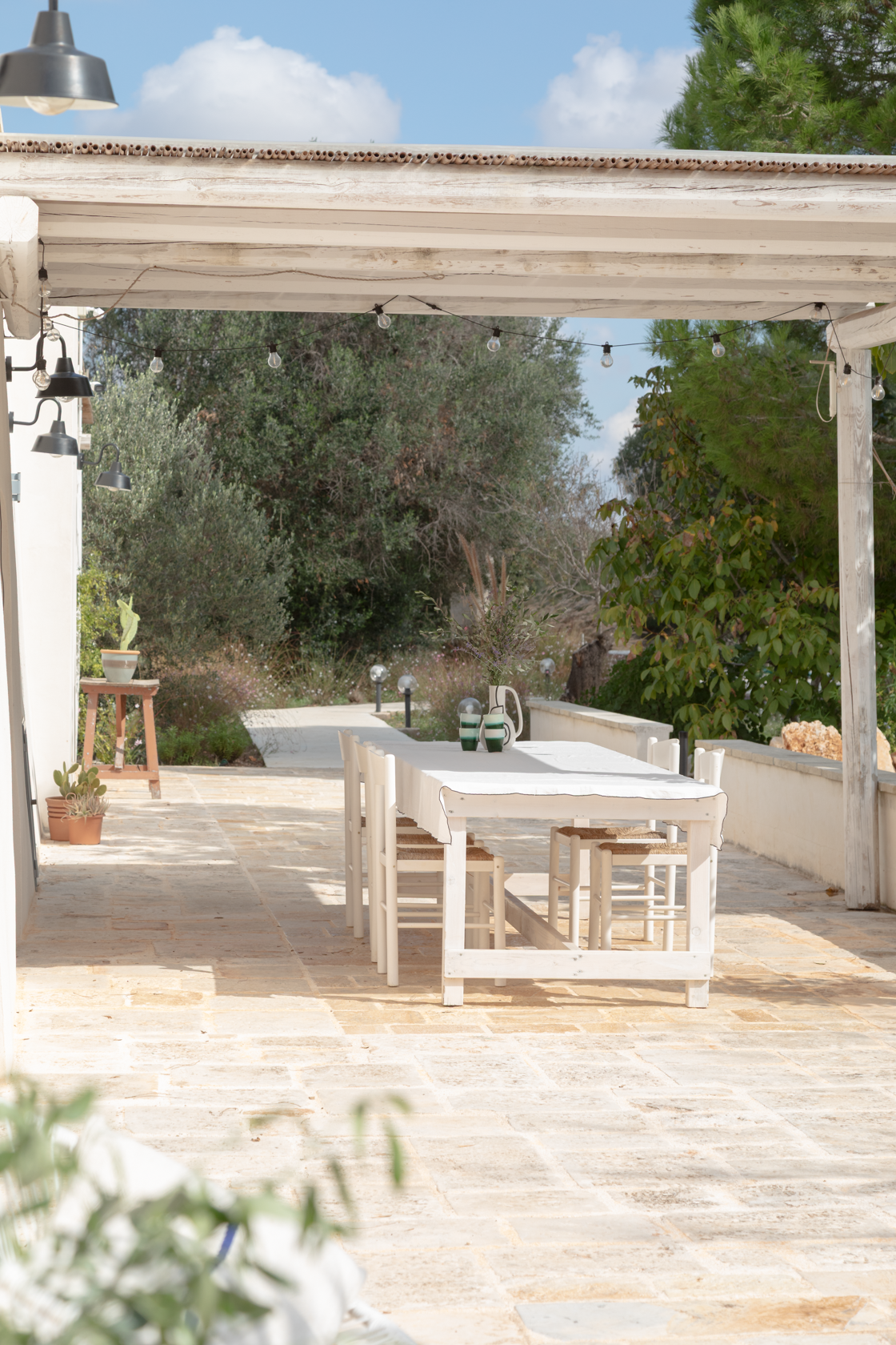 A white outdoor dining table with matching chairs under a pergola, decorated with vases, on a stone patio surrounded by green trees and plants.