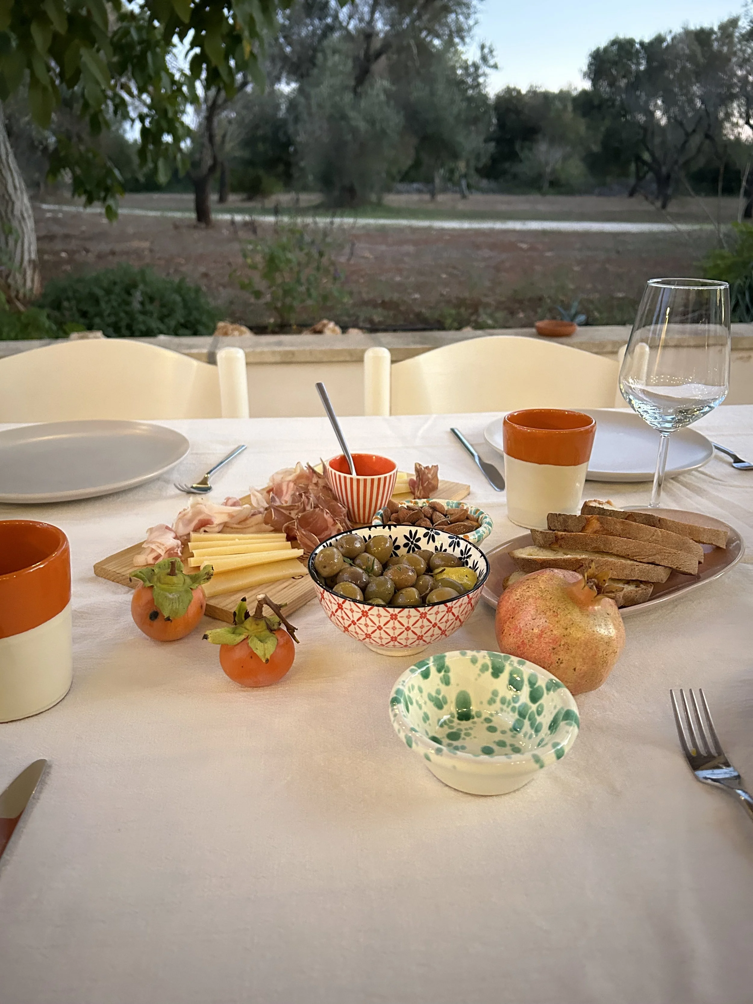A table set for a meal with olives, pomegranate, sliced bread, cheese, ham, and small bowls of condiments, with a scenic outdoor background.