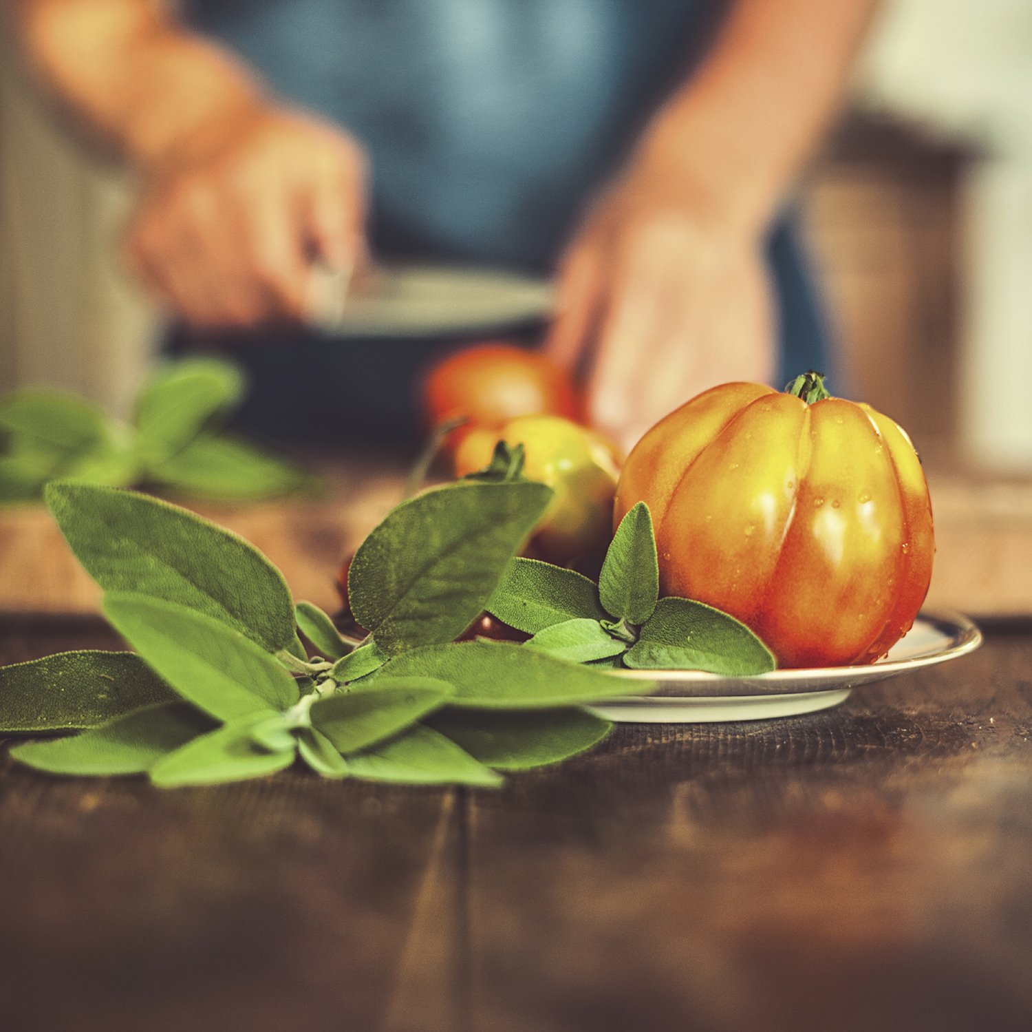 Close-up of heirloom tomatoes and green leaves on a plate, with a person slicing tomatoes in the background.