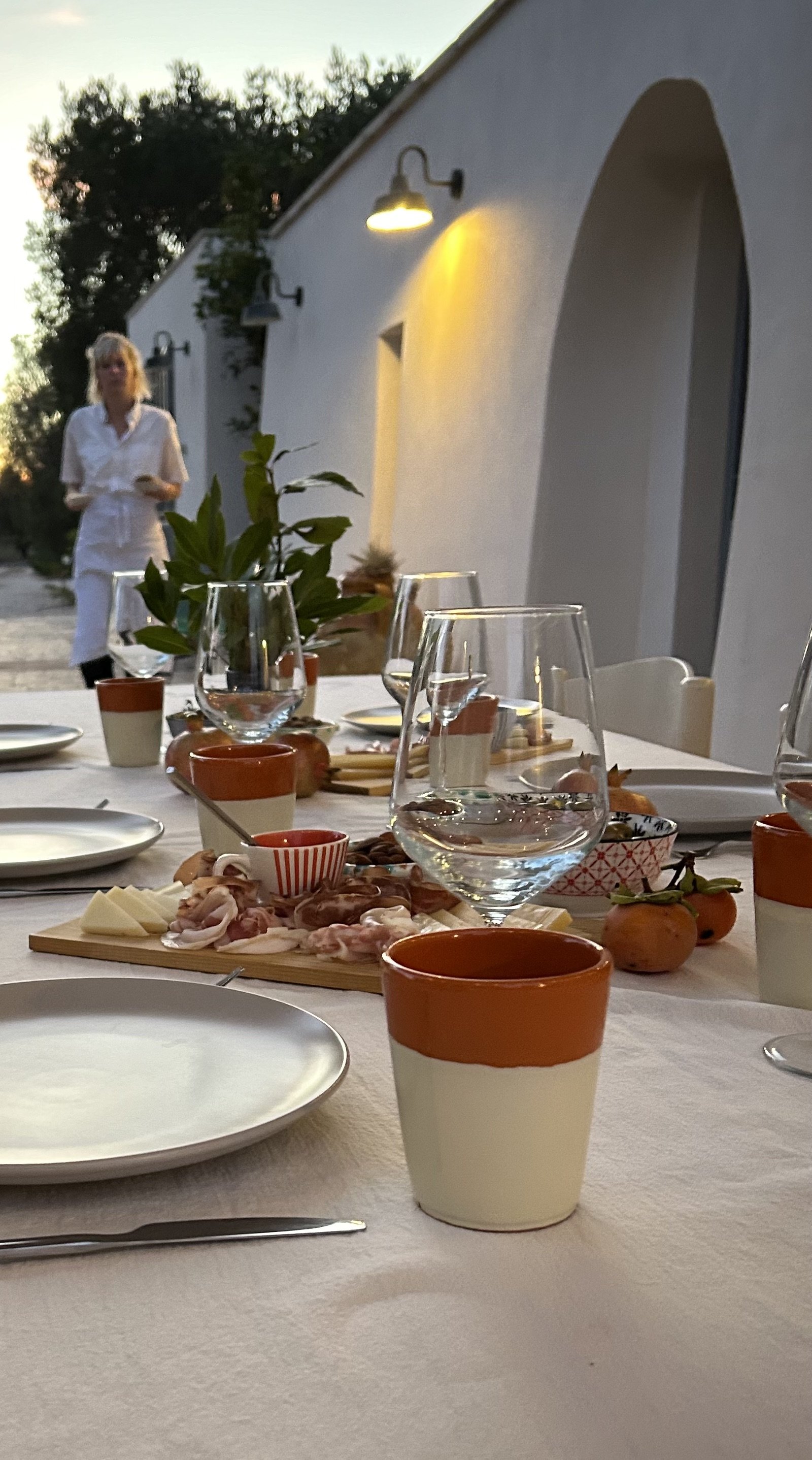 Table set for dinner outdoors with plates, glasses, and a charcuterie board, with a woman standing in the background near trullo fiorone illuminated by lights at sunset.