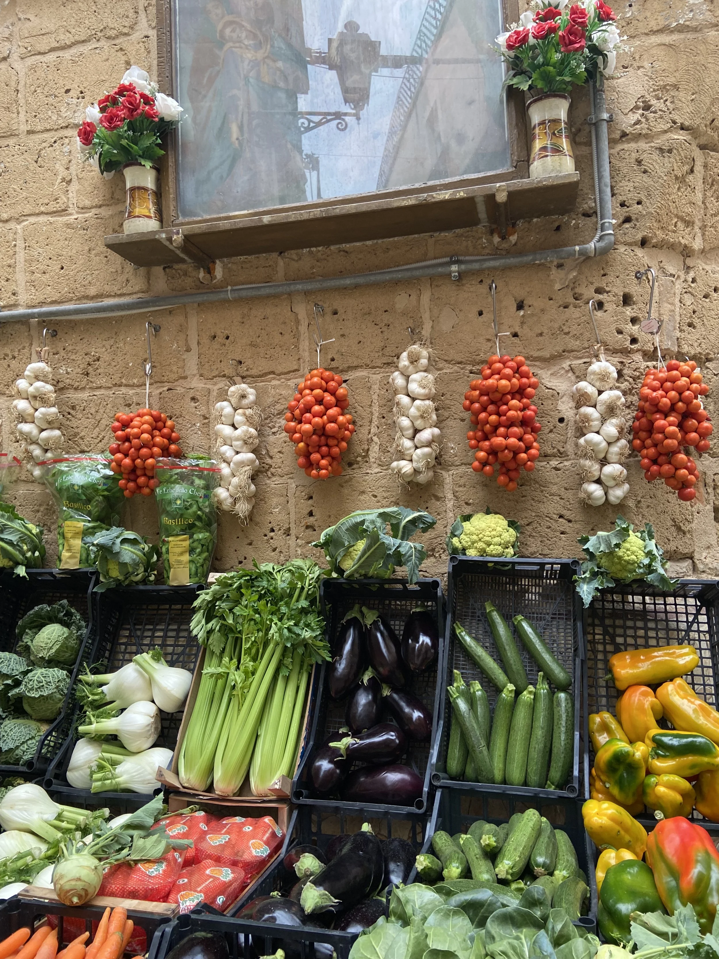 Fresh vegetables and herbs displayed on a market stand, including garlic, zucchinis, bell peppers, eggplant, carrots, and leafy greens, with dried tomatoes and garlic hanging above.