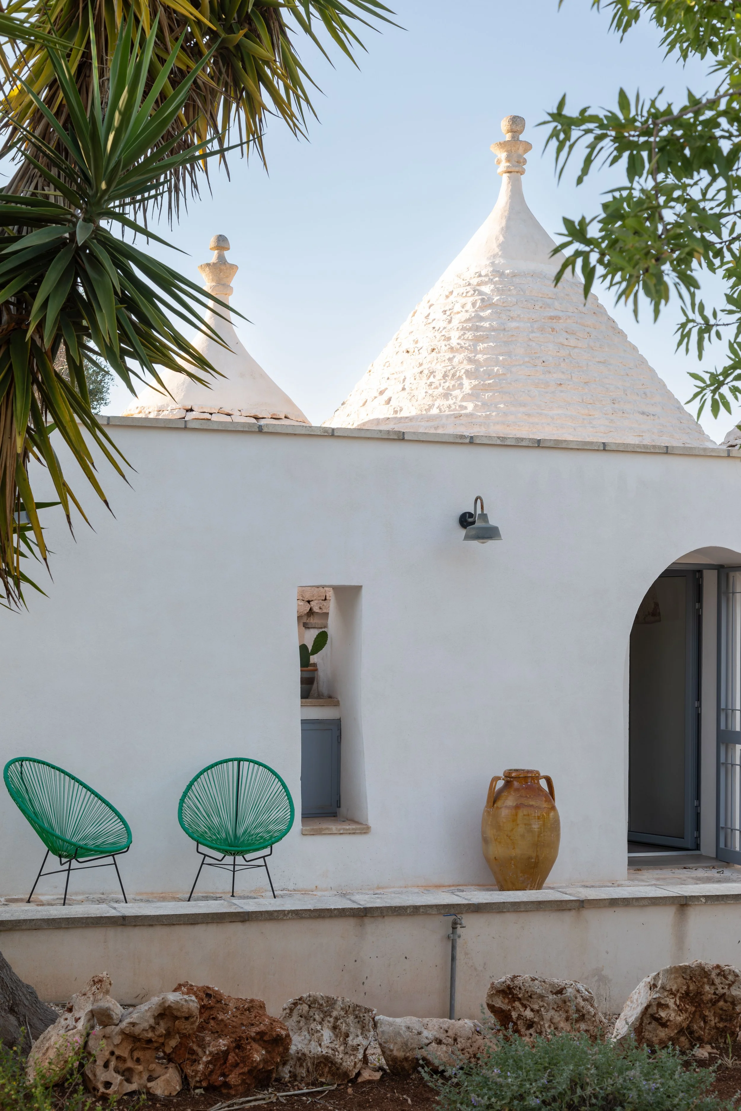 A white trullo with traditional conical roofs, two green wire chairs, a large clay pot, and an arched doorway, surrounded by desert plants and rocks.