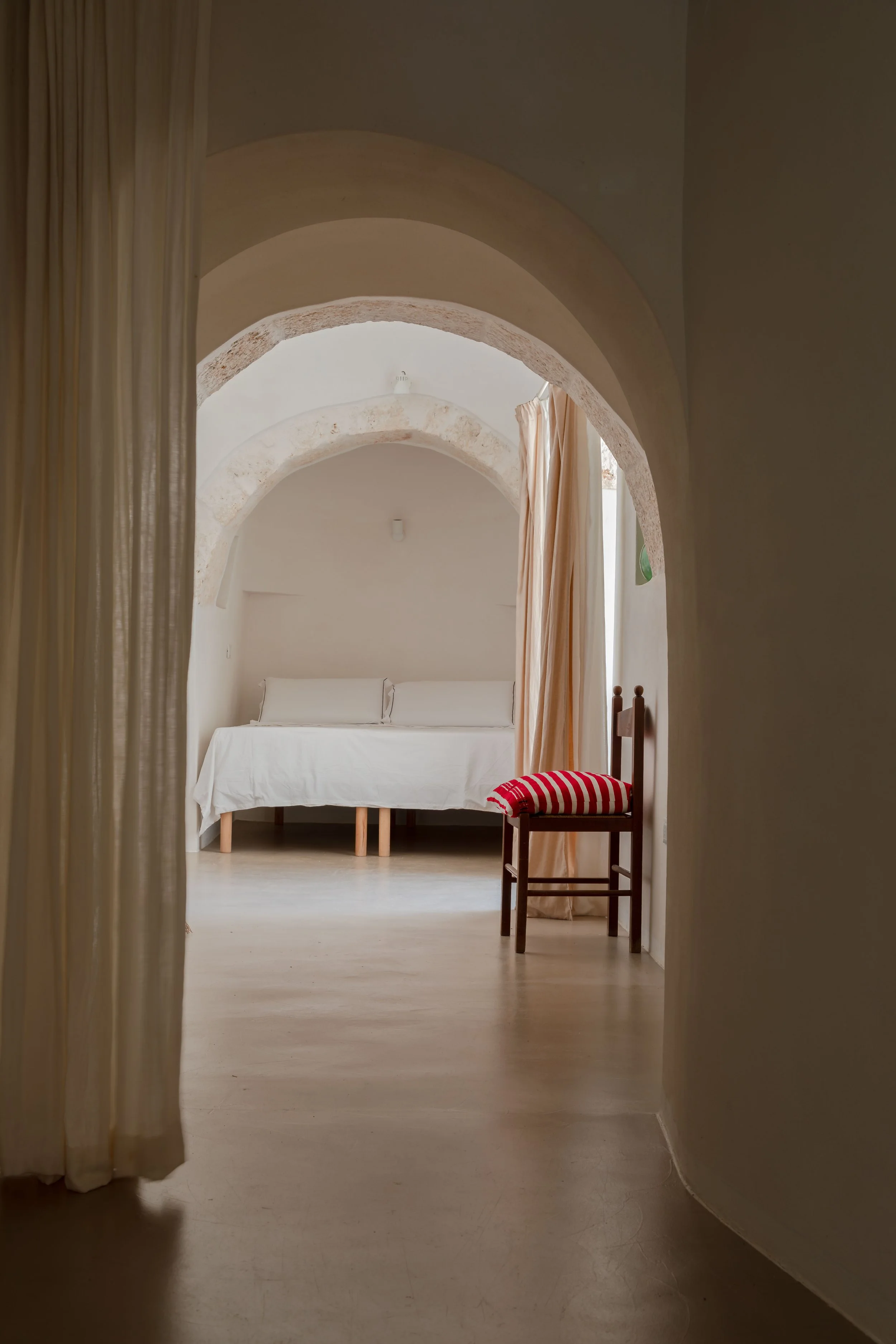 View of a minimalist bedroom with a bed, a chair with striped cushion, and arched doorways in neutral tones.