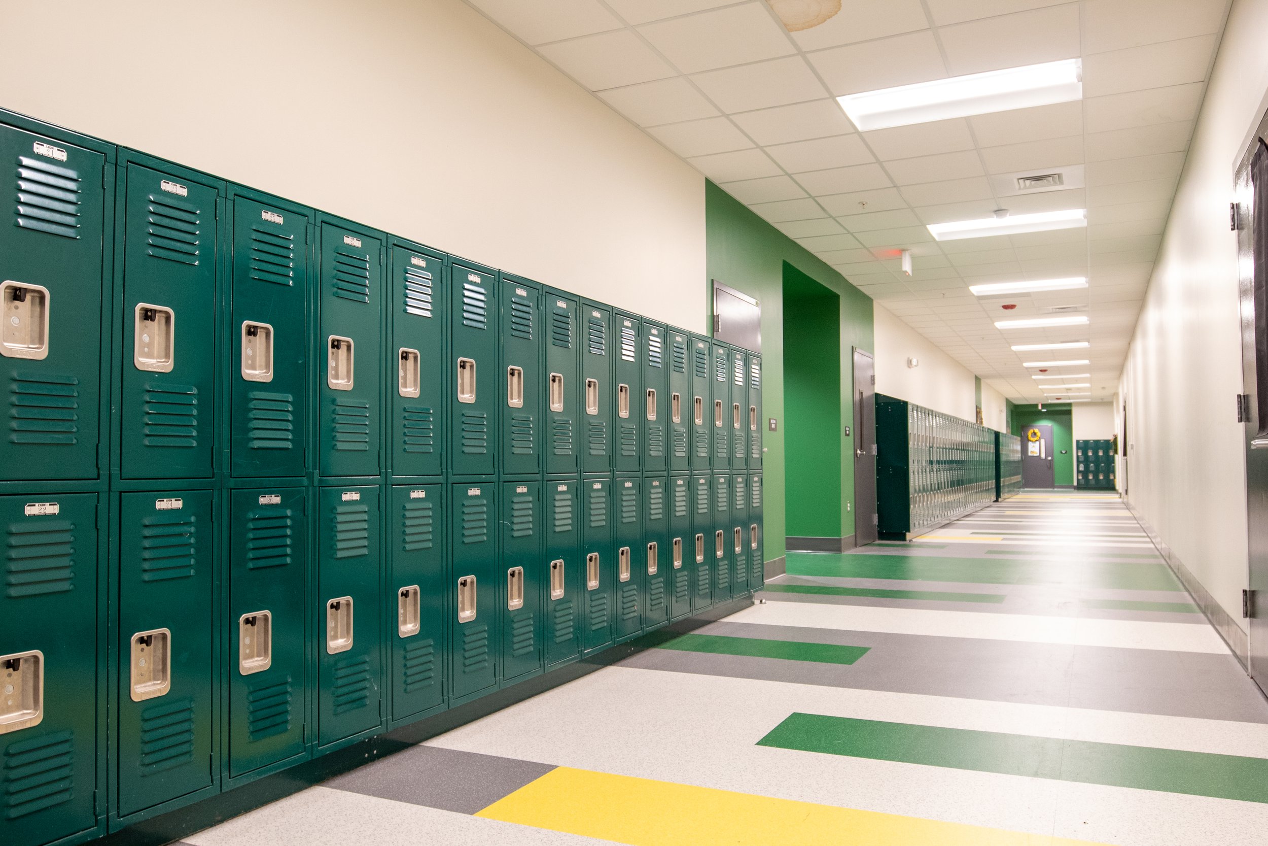 School hallway with green lockers on the left and lockers in the distance on the right, with a light-colored tiled floor and a drop ceiling.