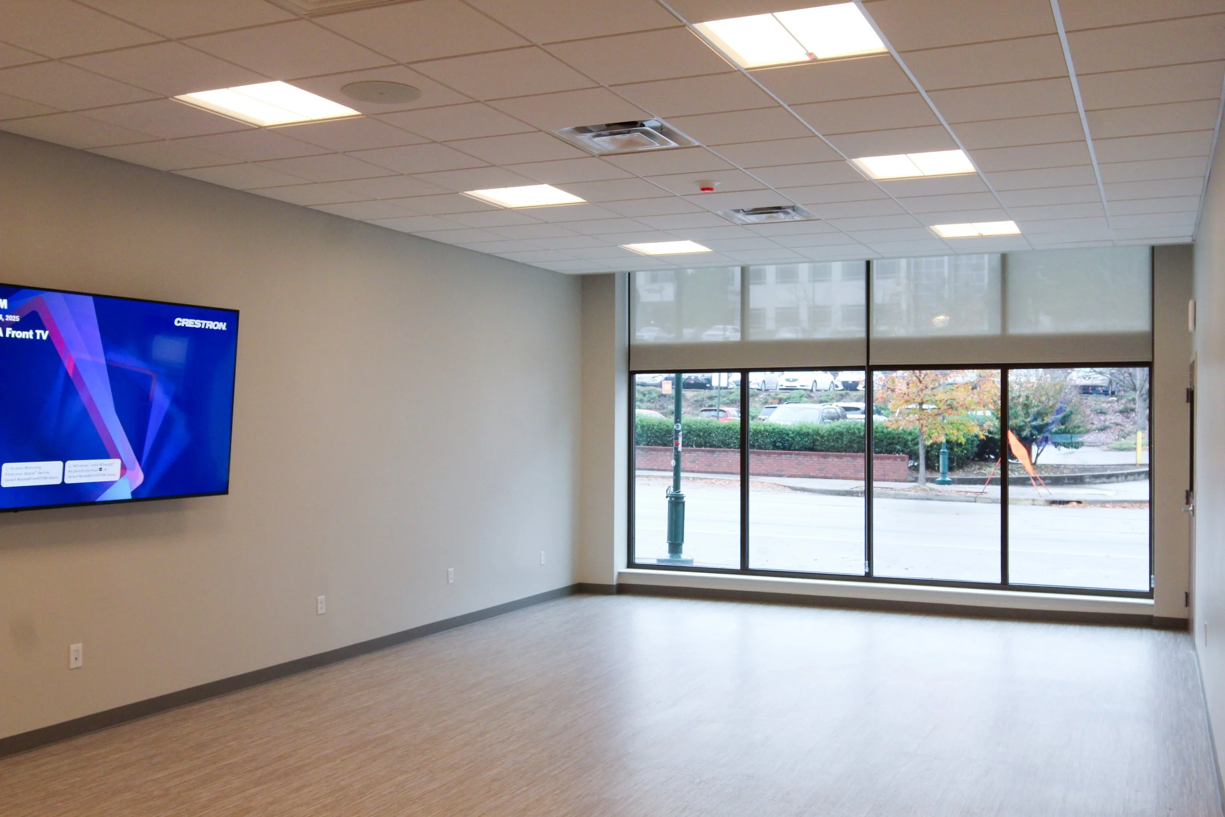 Empty office space with large front window, wall-mounted flat-screen TV on the left, neutral-colored walls, beige carpet, ceiling with recessed lighting and air vents, and a view of a parking lot and outdoor landscape through the window.