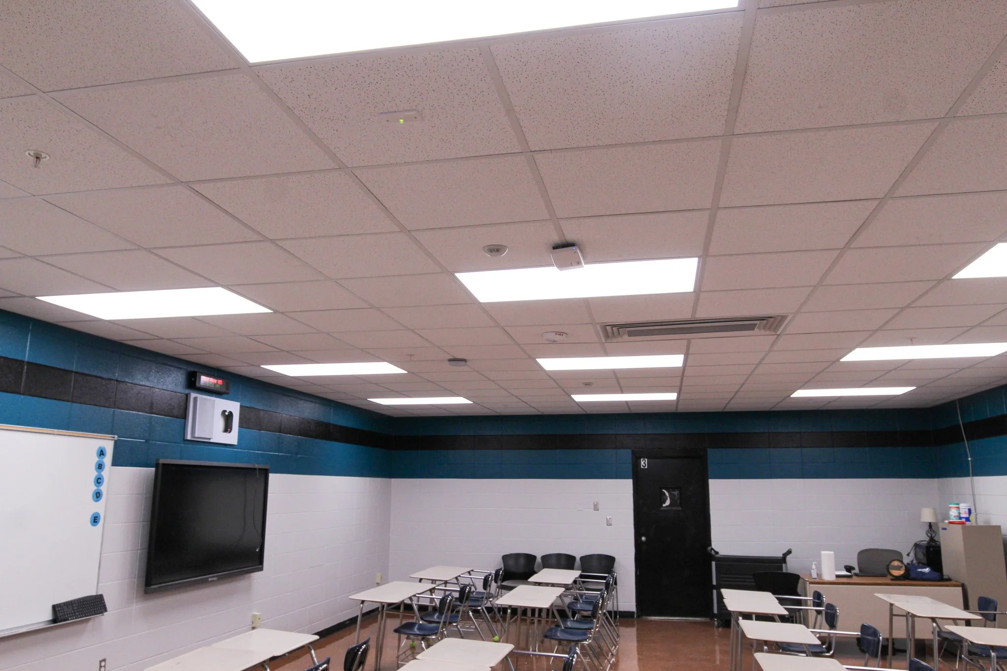 A classroom with white ceiling tiles, some fluorescent lights, and a blue wall. There is a television mounted on the white wall, and several desks and chairs are arranged in the room. The room appears to be part of a school or educational facility.