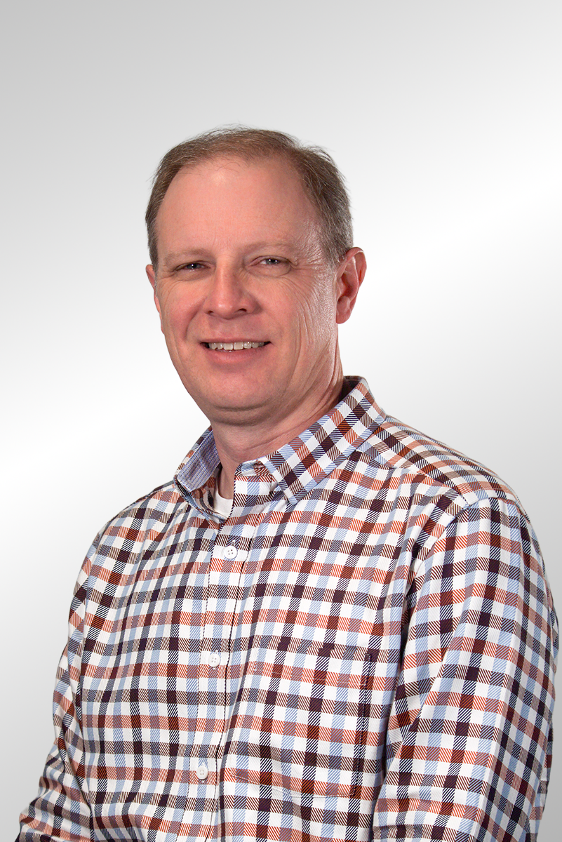 A man with short reddish hair and fair skin wearing a red, tan, and blue plaid button-up shirt, standing against a dark gray background.