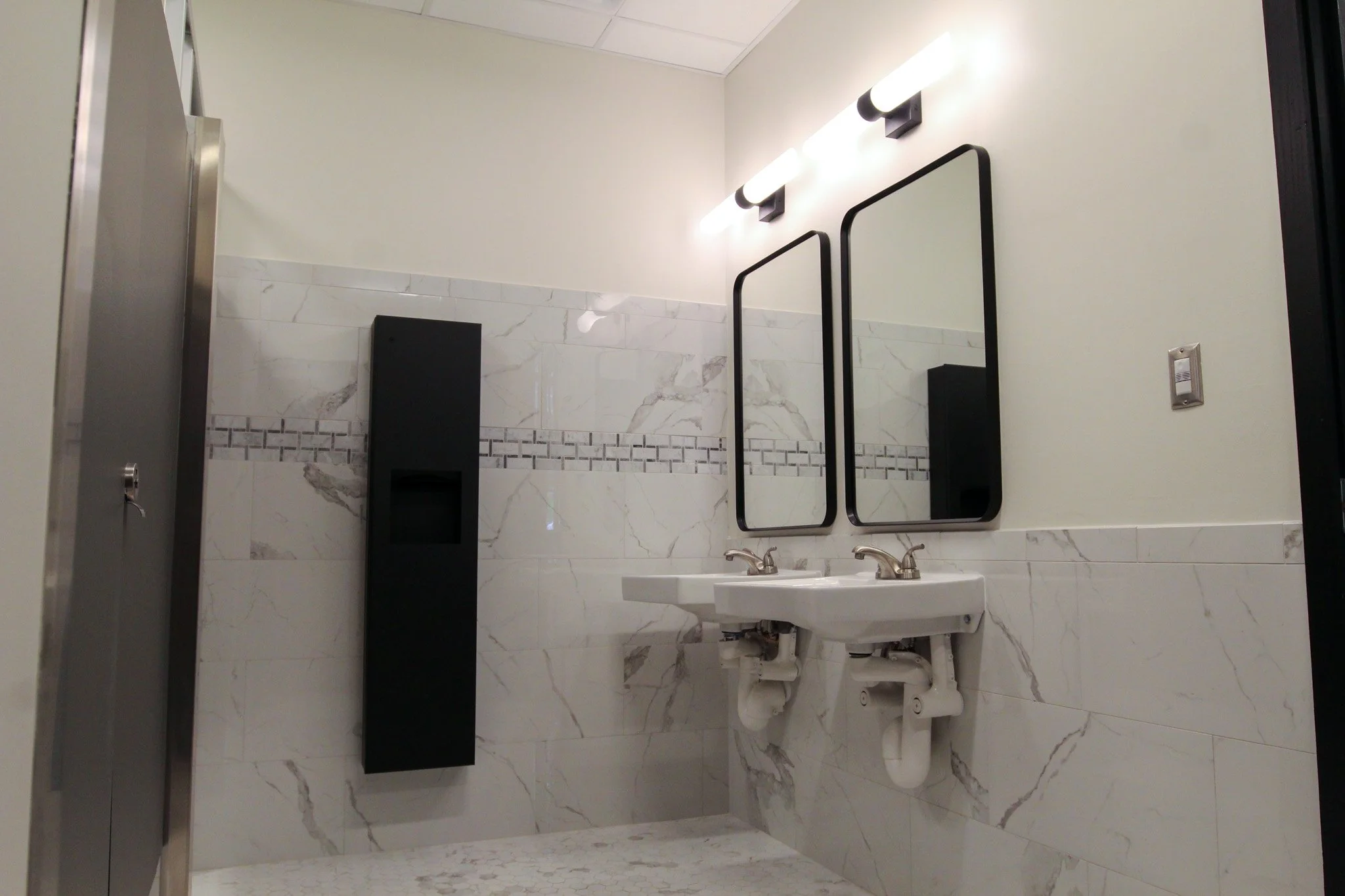 A public restroom with marble tile walls and floor, two sinks with faucets, rectangular mirrors, a soap dispenser, and black fixtures. The lighting is bright with two wall-mounted lights above the mirrors.