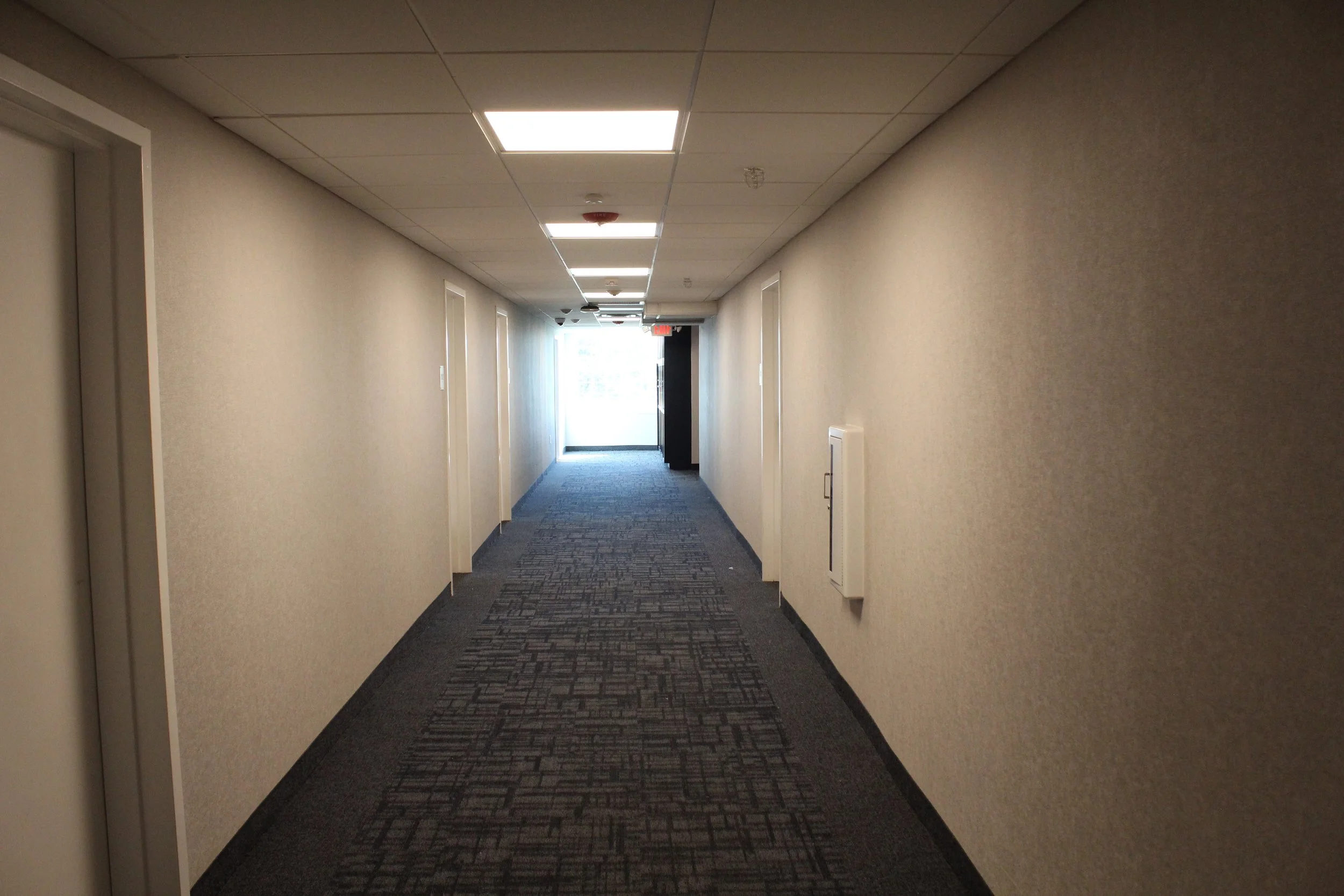 A long, empty hallway in a building with beige walls, carpeted flooring, and fluorescent ceiling lights. Doors are visible on both sides, and an exit door with a window is at the end of the hallway.