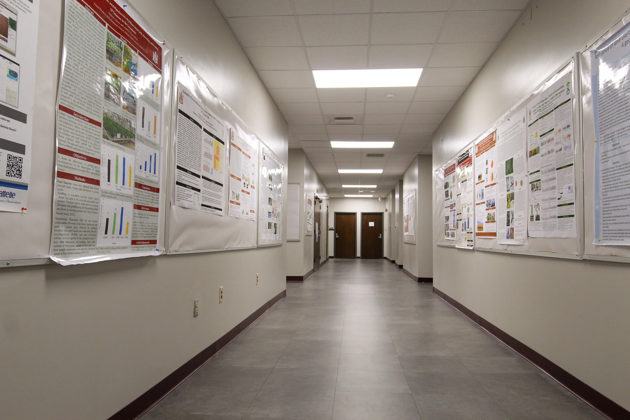 Empty hallway with white walls and bulletin boards on both sides displaying various science research posters, leading to a double door at the end.